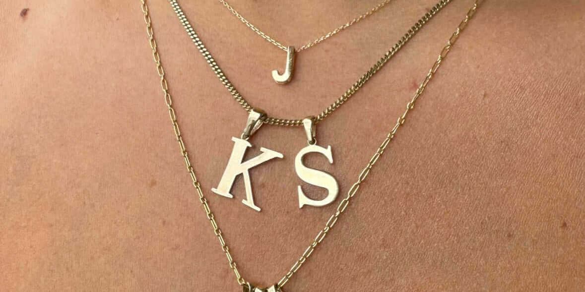 Close-up of layered gold personalized initials jewelry featuring 'J', 'KS', and charm necklaces.