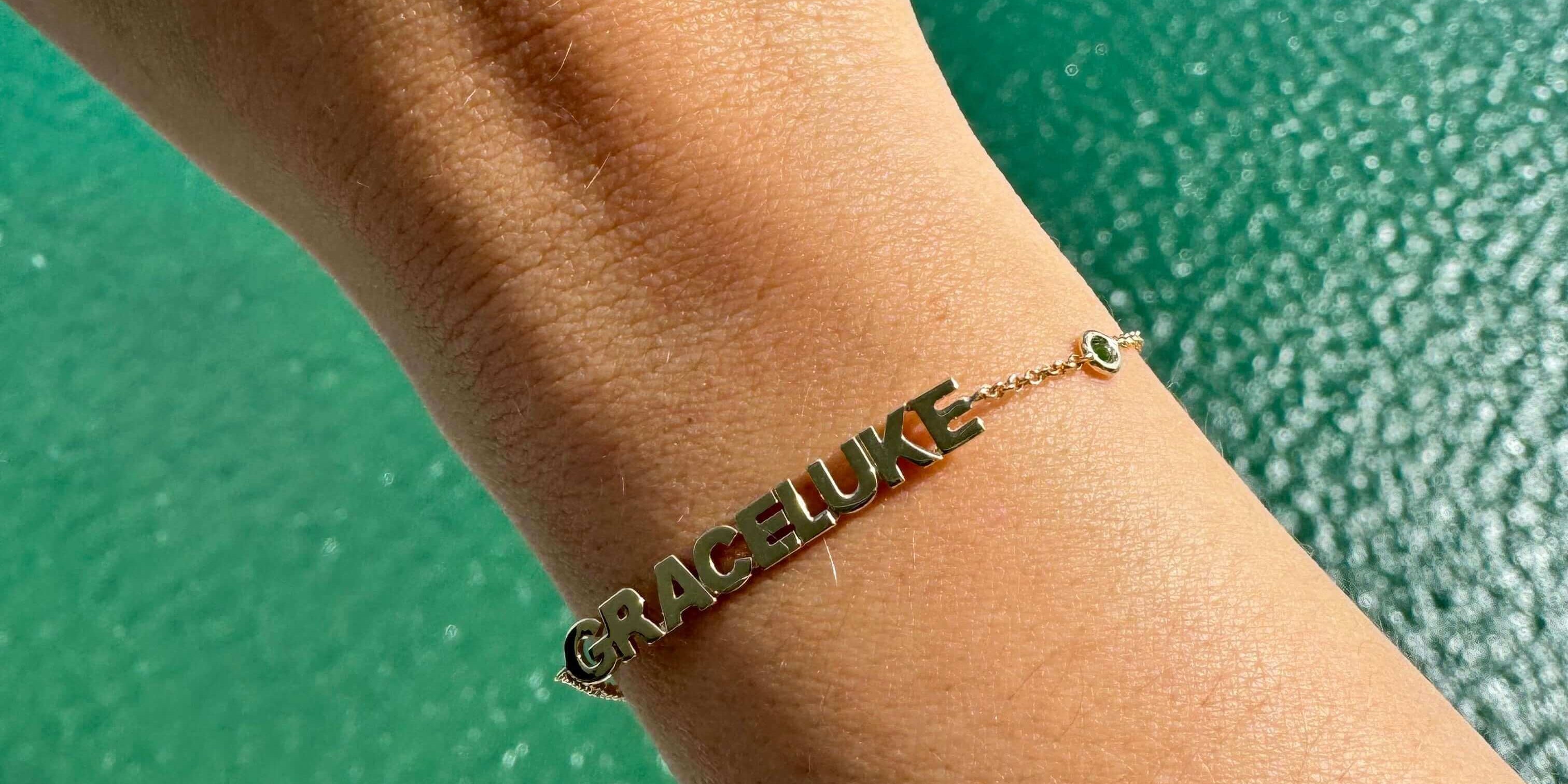 Personalized gold bracelet with names 'Grace' and 'Luke' displayed, set against a turquoise water background.