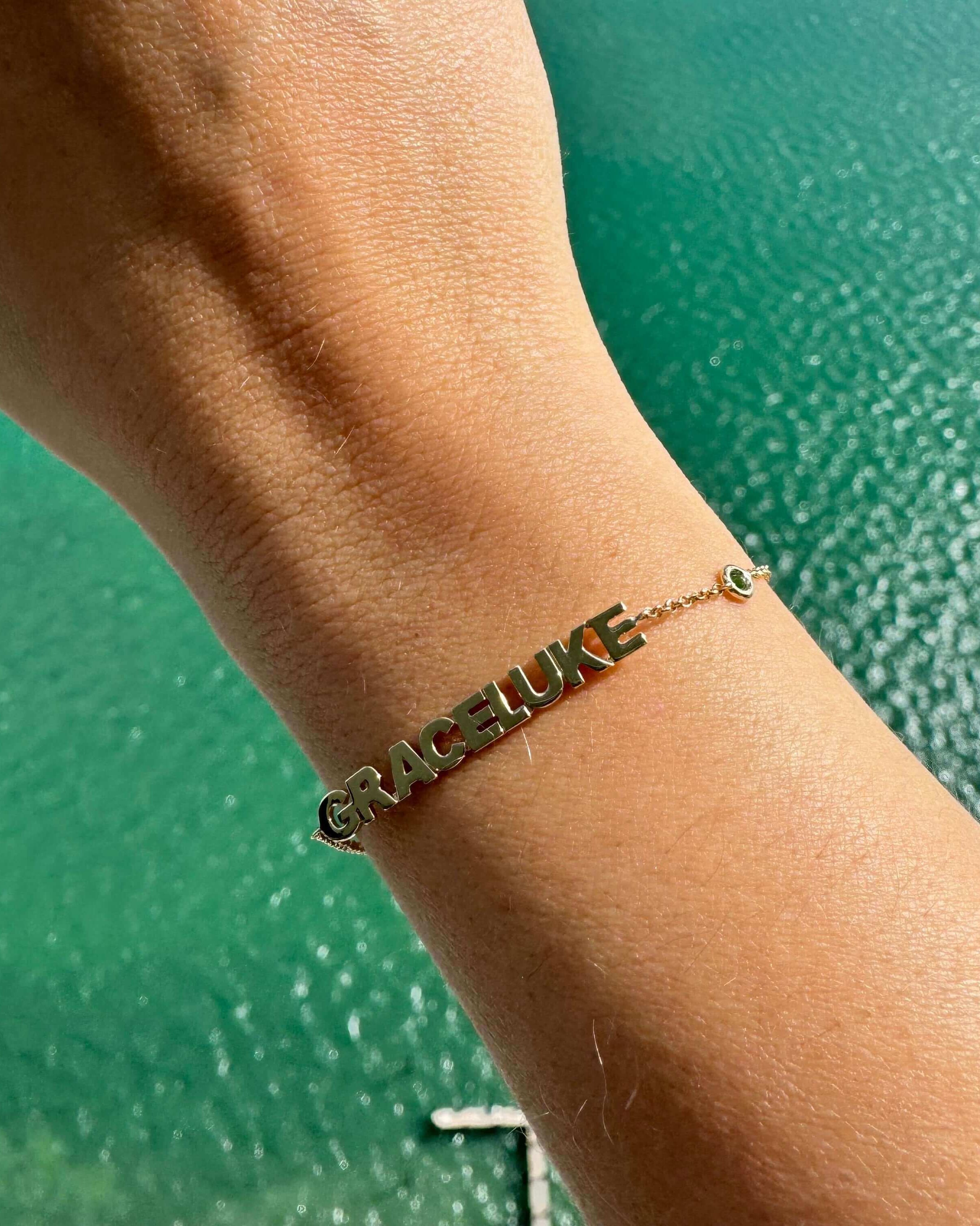 Personalized gold bracelet with names 'Grace' and 'Luke' displayed, set against a turquoise water background.