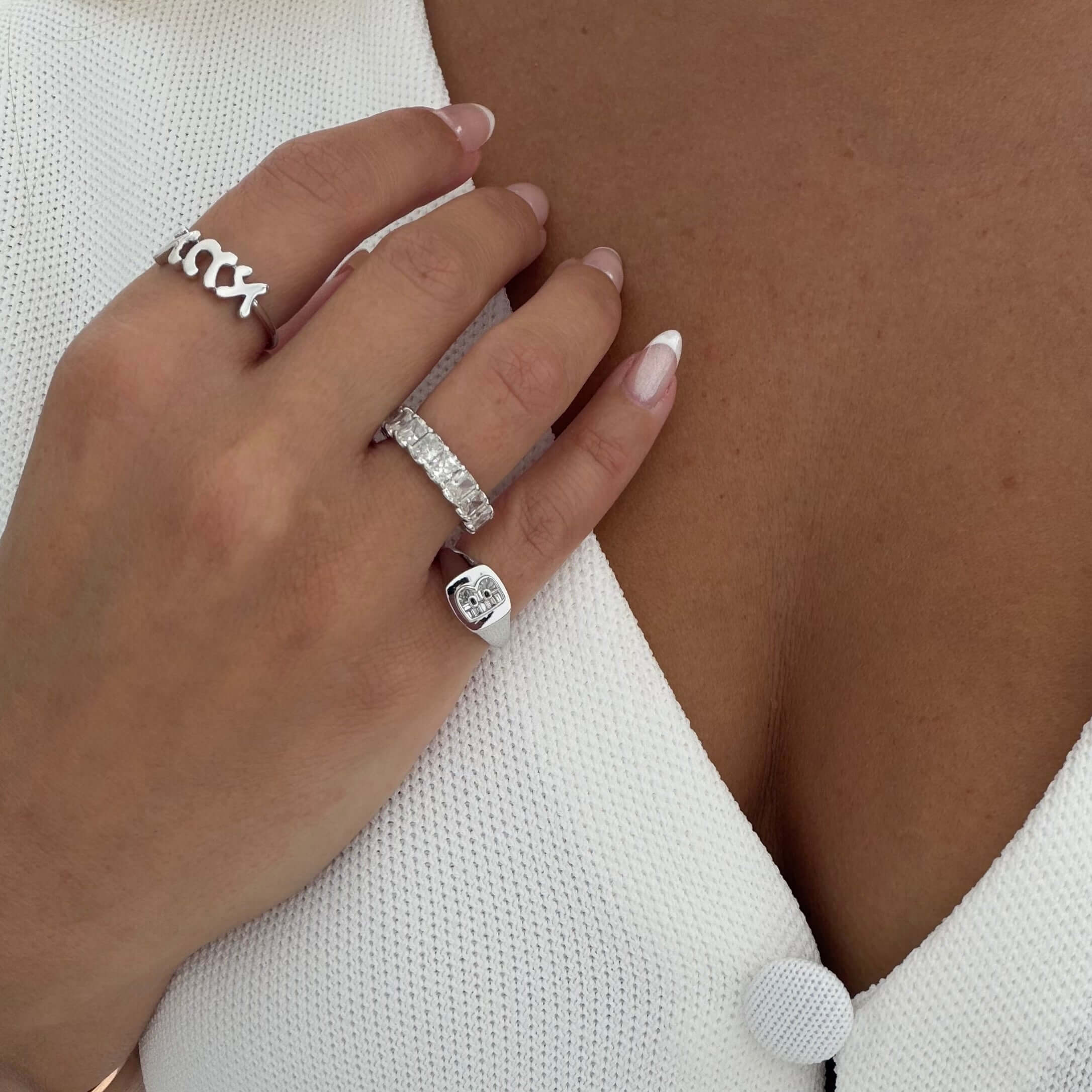 Woman's hand displaying multiple rings, including a Baguette Diamond Initial Signet Ring, against a textured white background.
