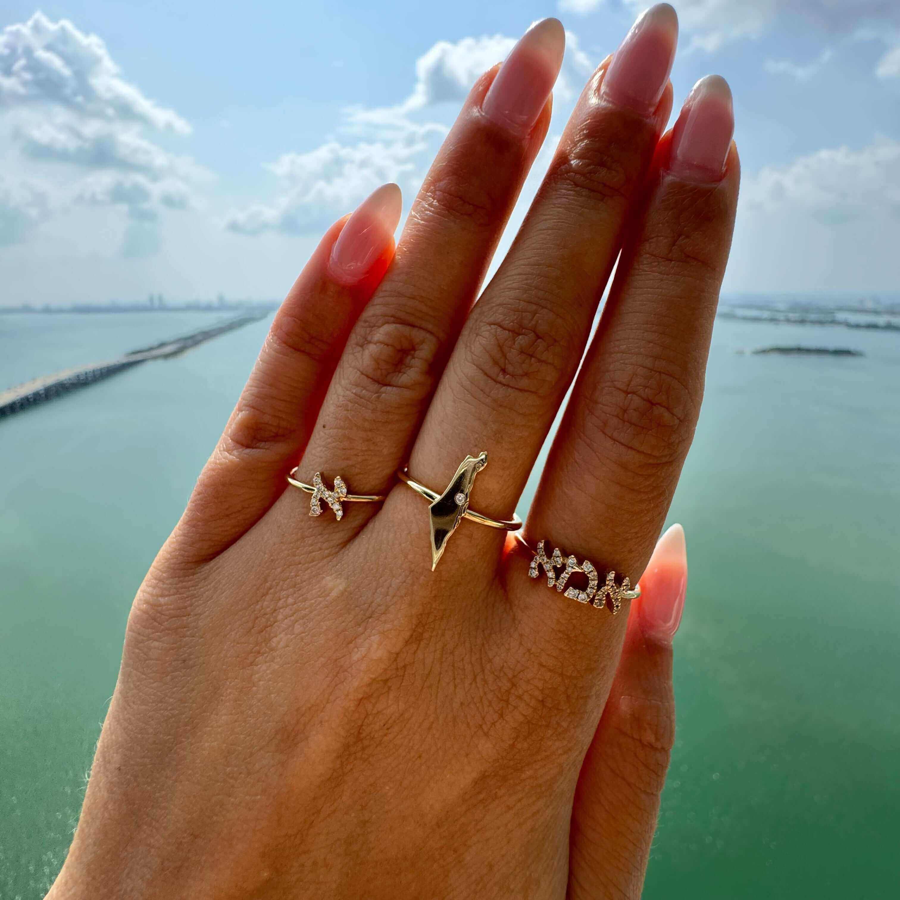 Hand adorned with gold and diamond rings overlooking serene body of water, showcasing our luxury jewelry brand.