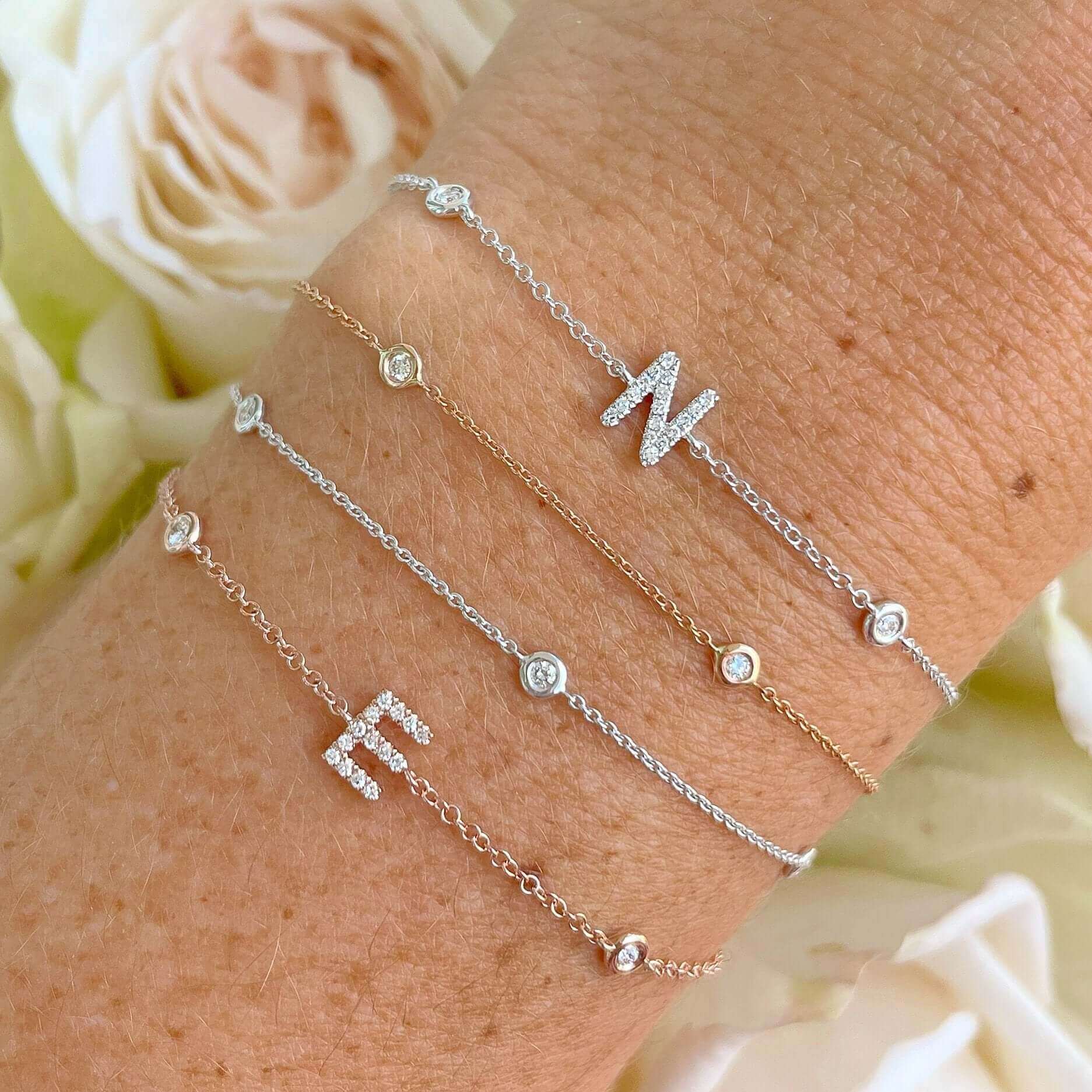 Elegant initial charm bracelets in gold and silver with diamond accents on wrist, showcasing delicate jewelry design against a background of white roses.