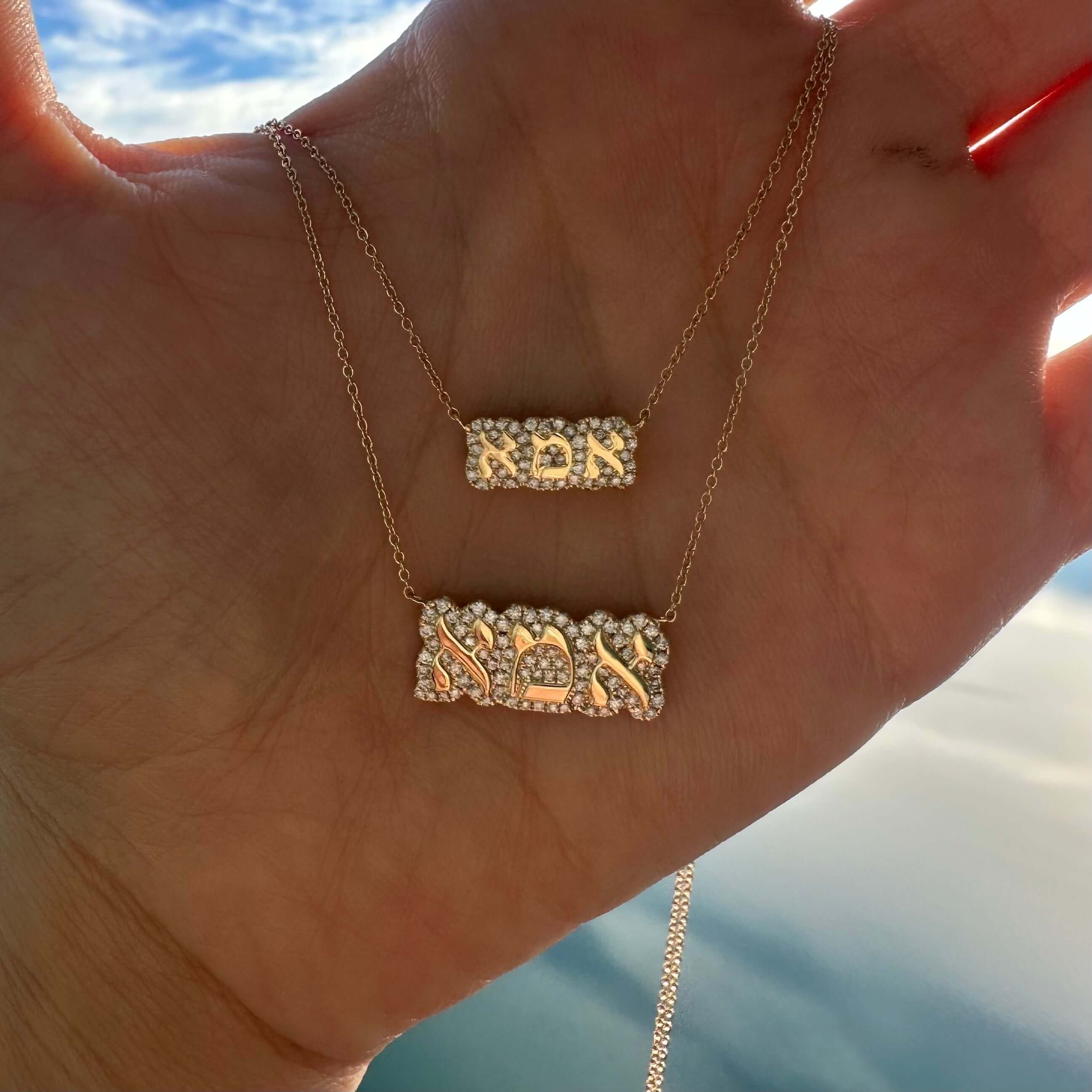 Double-layered necklace with intricate Hebrew pendant design held against a scenic sky backdrop by a hand.