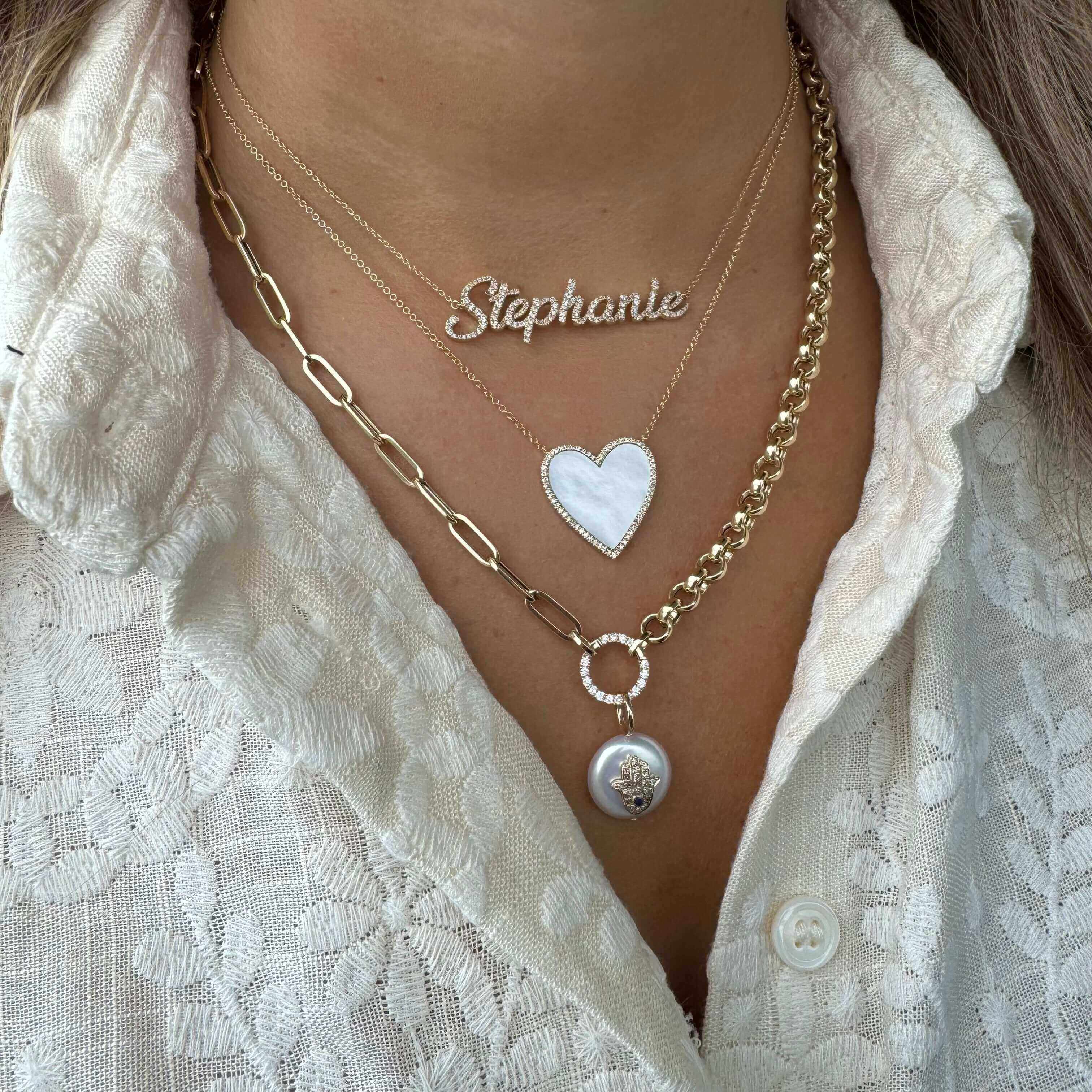 Layered necklaces featuring a custom nameplate, heart pendant, and chain link design on a textured white blouse.