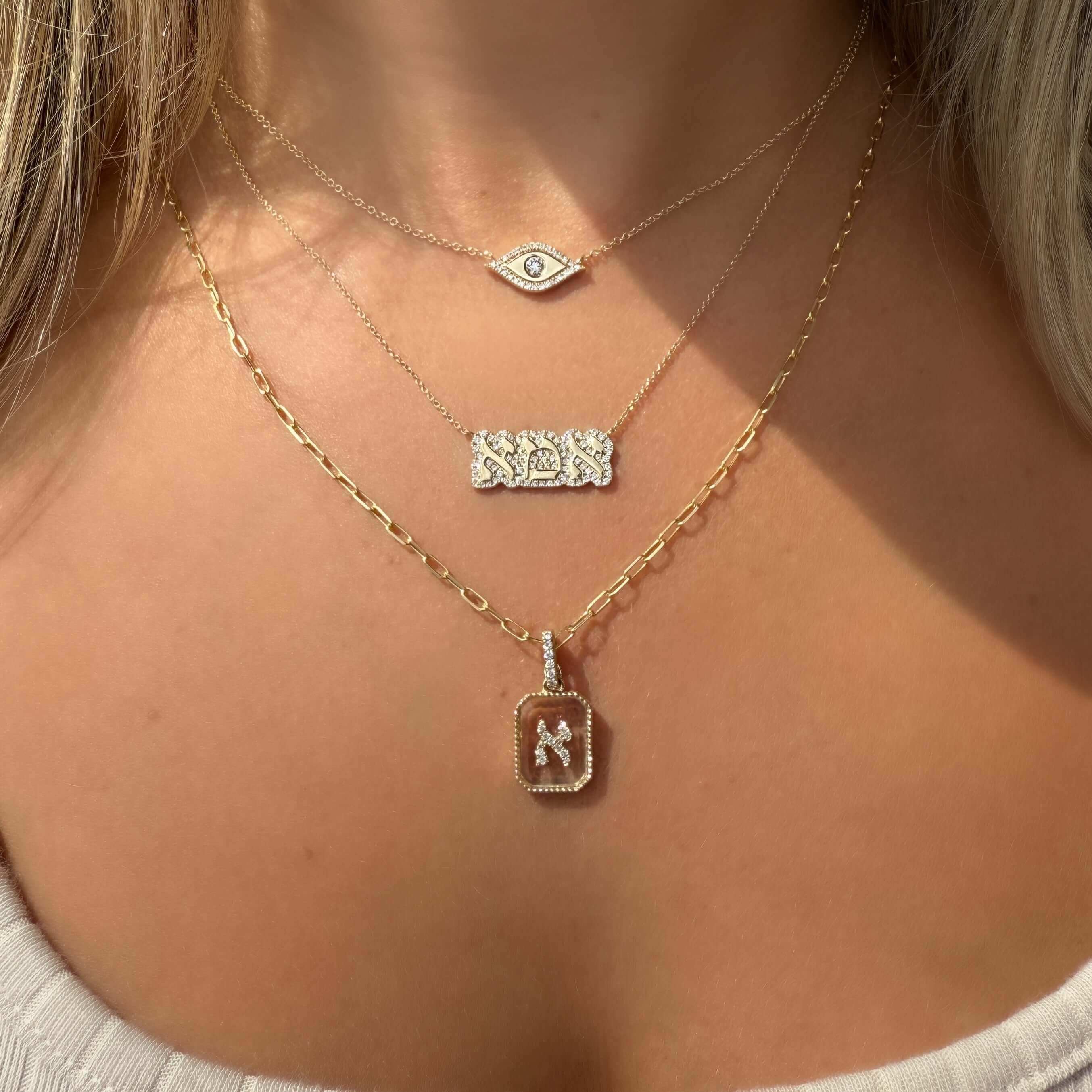 Model wearing layered necklaces featuring a diamond Hebrew initial pendant, an eye symbol, and intricate designs.