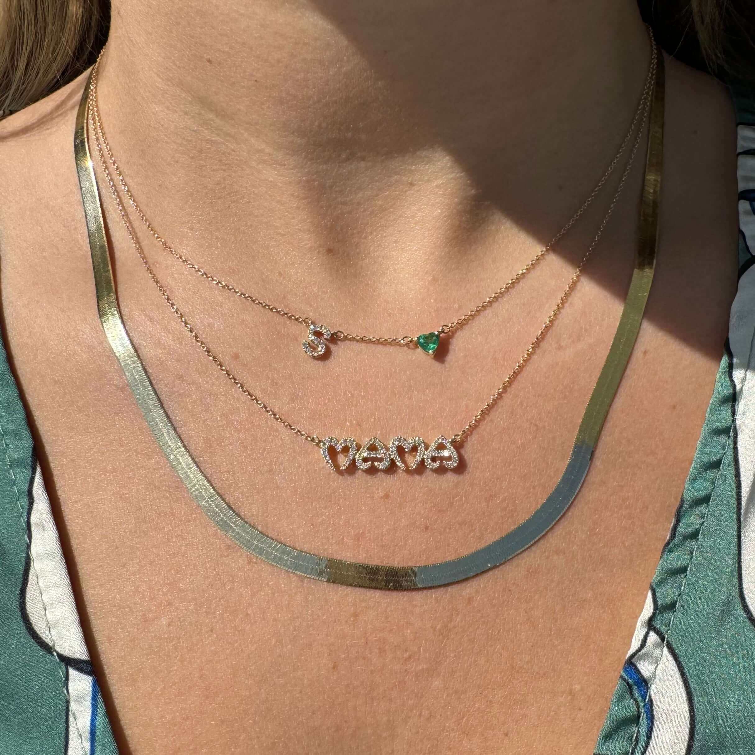 Woman wearing layered necklaces from our jewelry brand featuring a silver herringbone chain, an initial 'S' charm, and a green gemstone pendant.