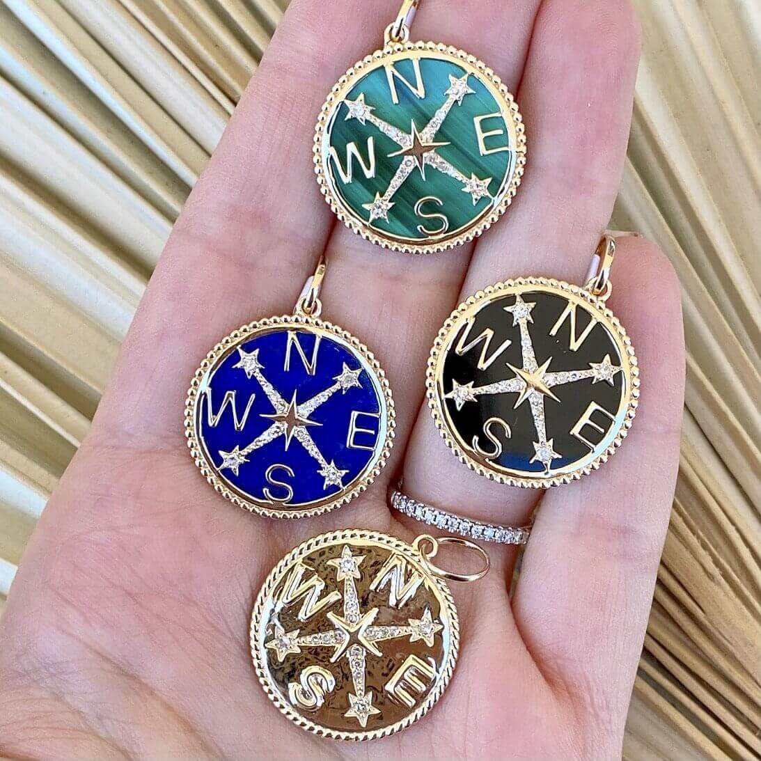 Hand holding four compass-themed pendants in green, blue, black, and gold from our jewelry collection