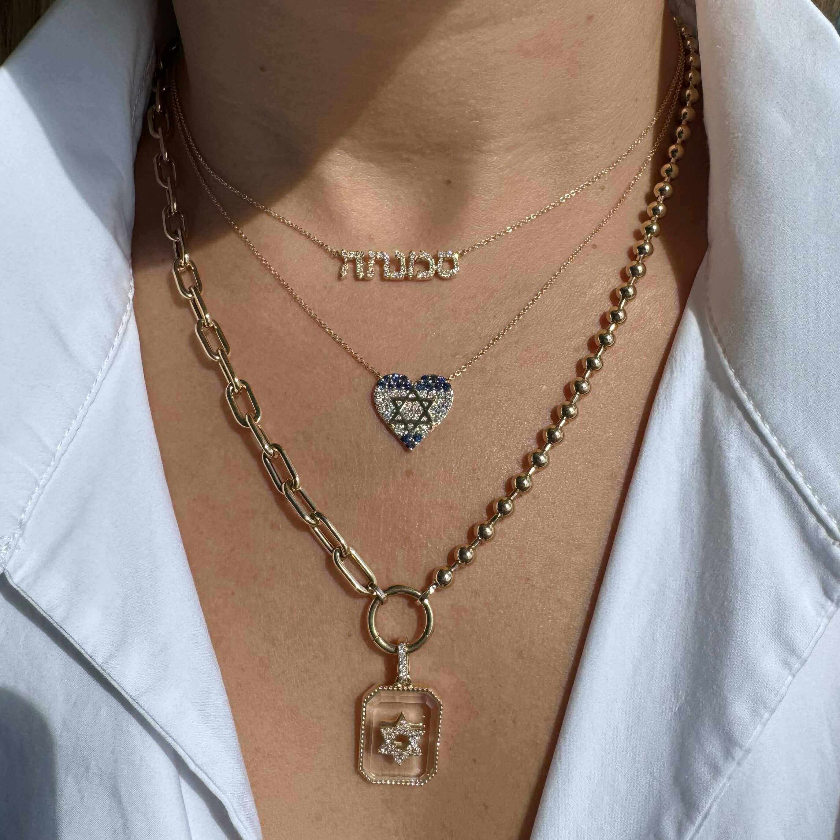 Model showcasing layered gold necklaces with heart, charm, and paper clip chain designs against a plain white shirt.