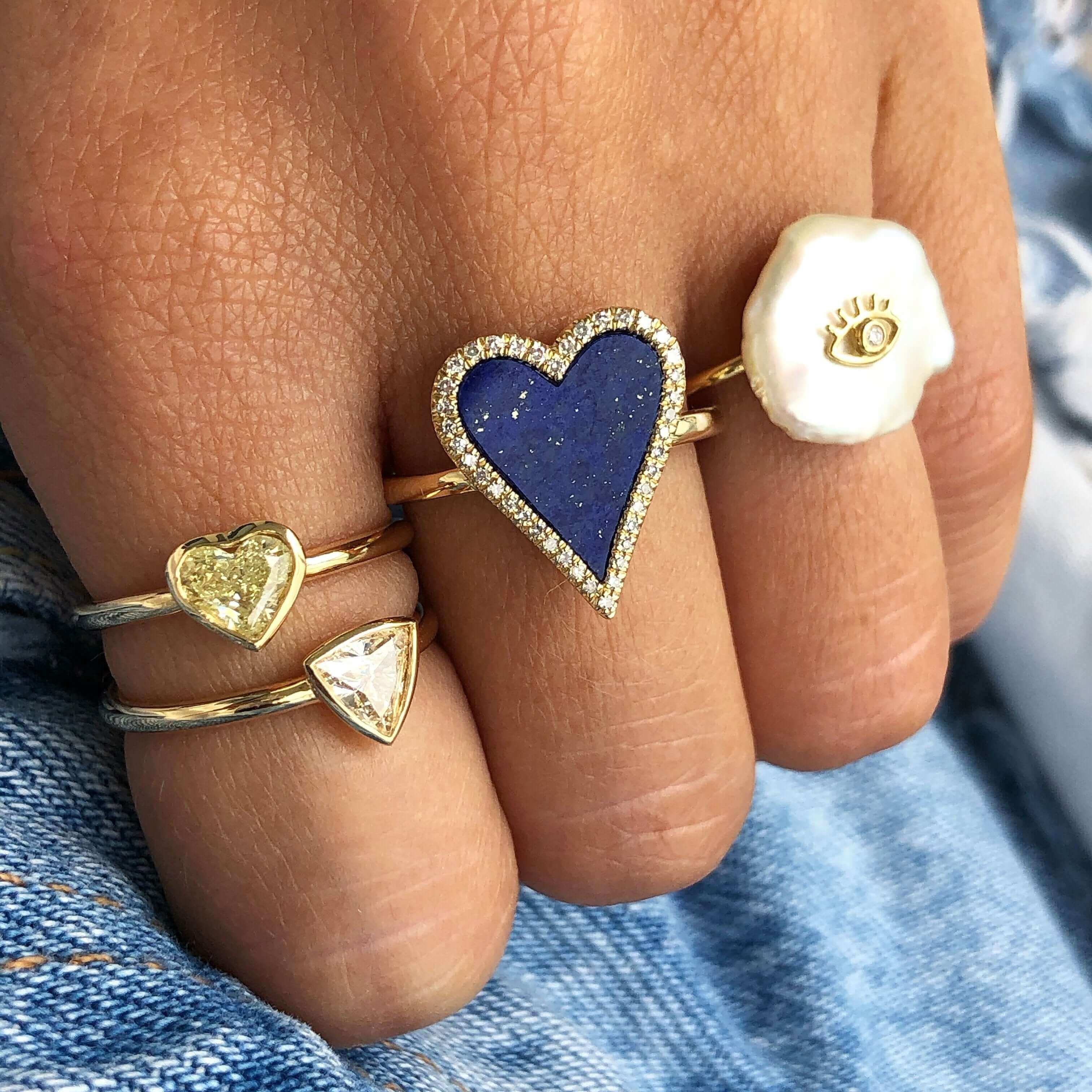 Hand wearing trendy heart-shaped and eye-design rings, showcasing our exclusive jewelry collection against a denim backdrop.