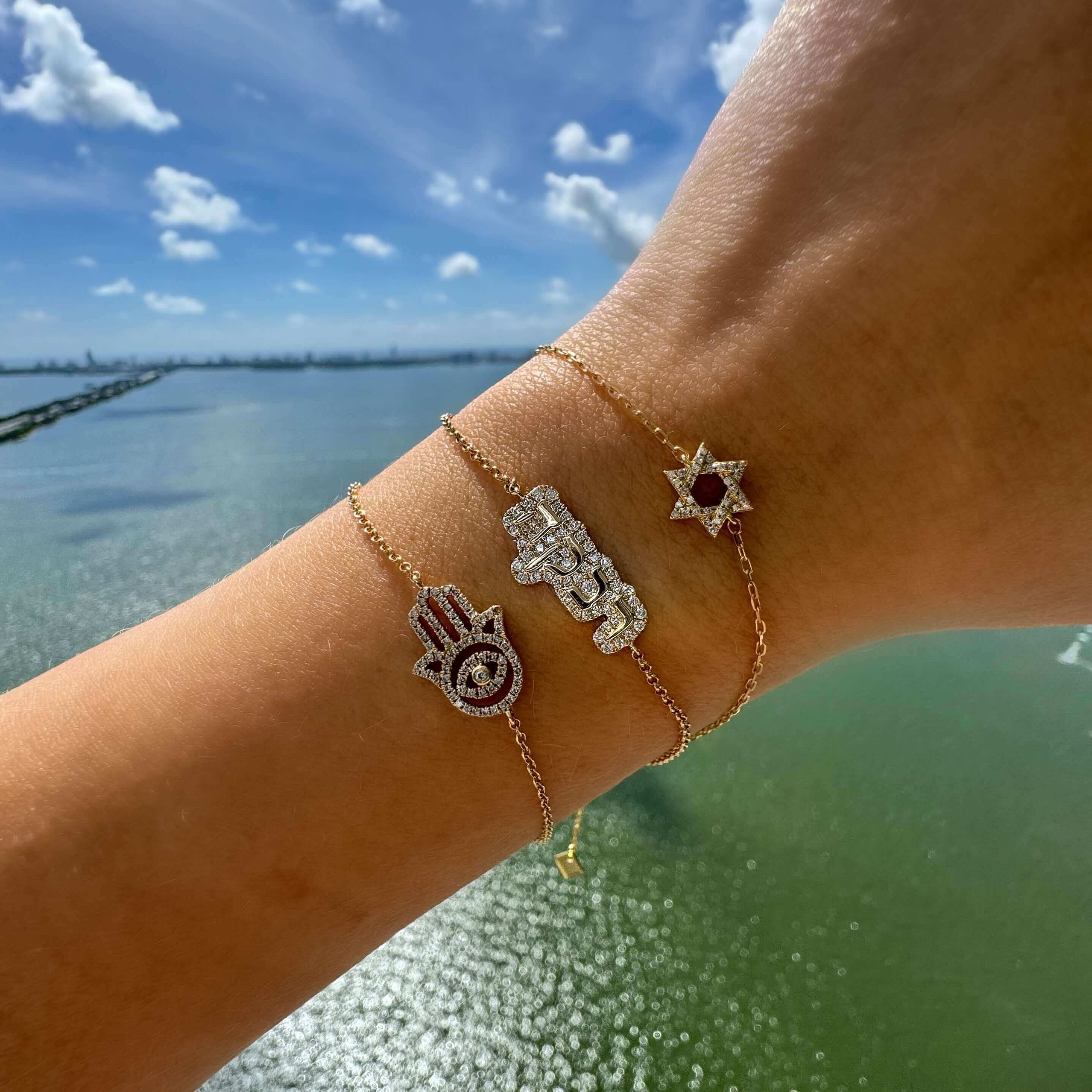 Gold bracelet collection featuring Hamsa Hand, Hebrew symbol, and Star of David charms overlooking a serene waterfront background.
