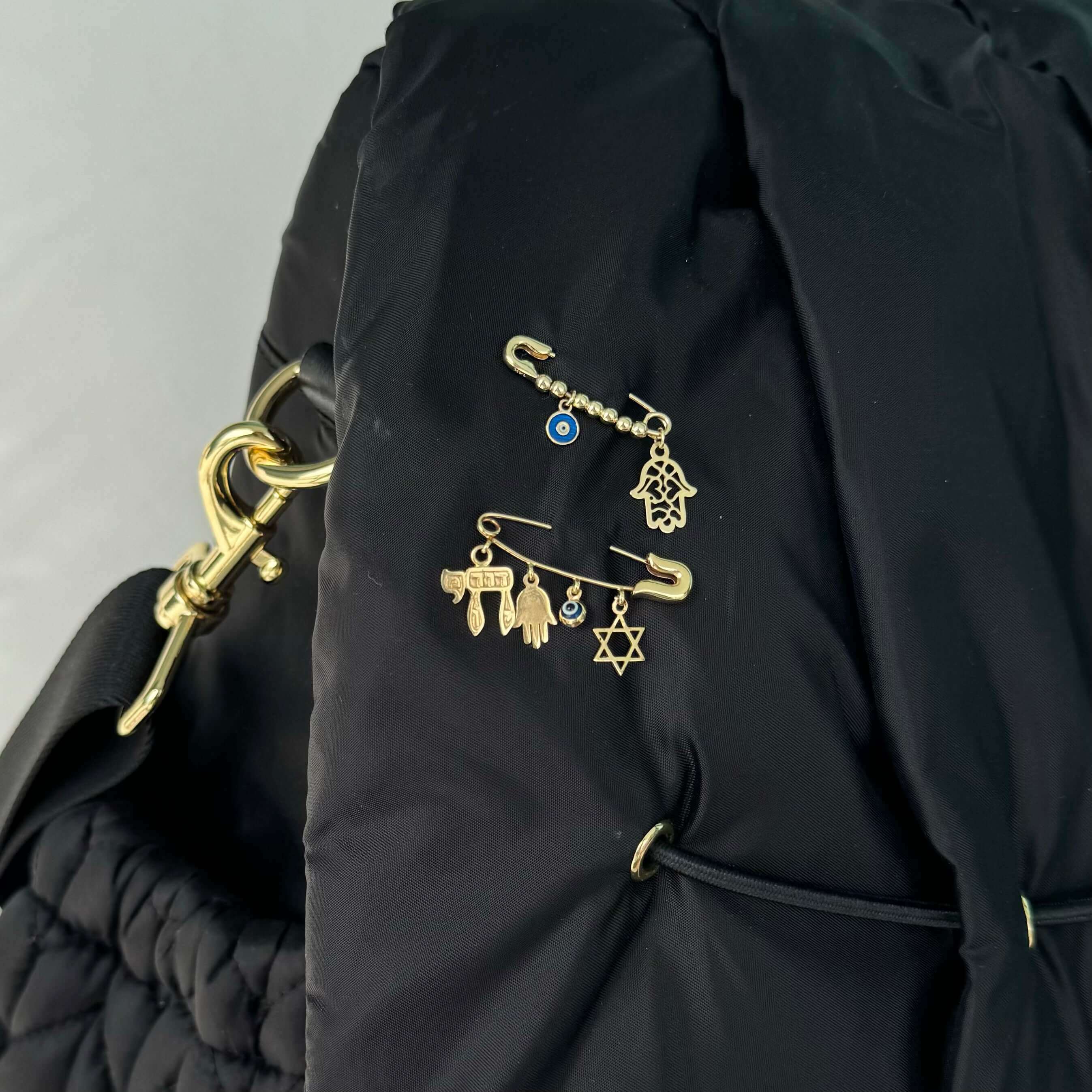 Gold charm pins featuring various designs including a star and hamsa hand, attached to a black quilted bag from our jewelry brand