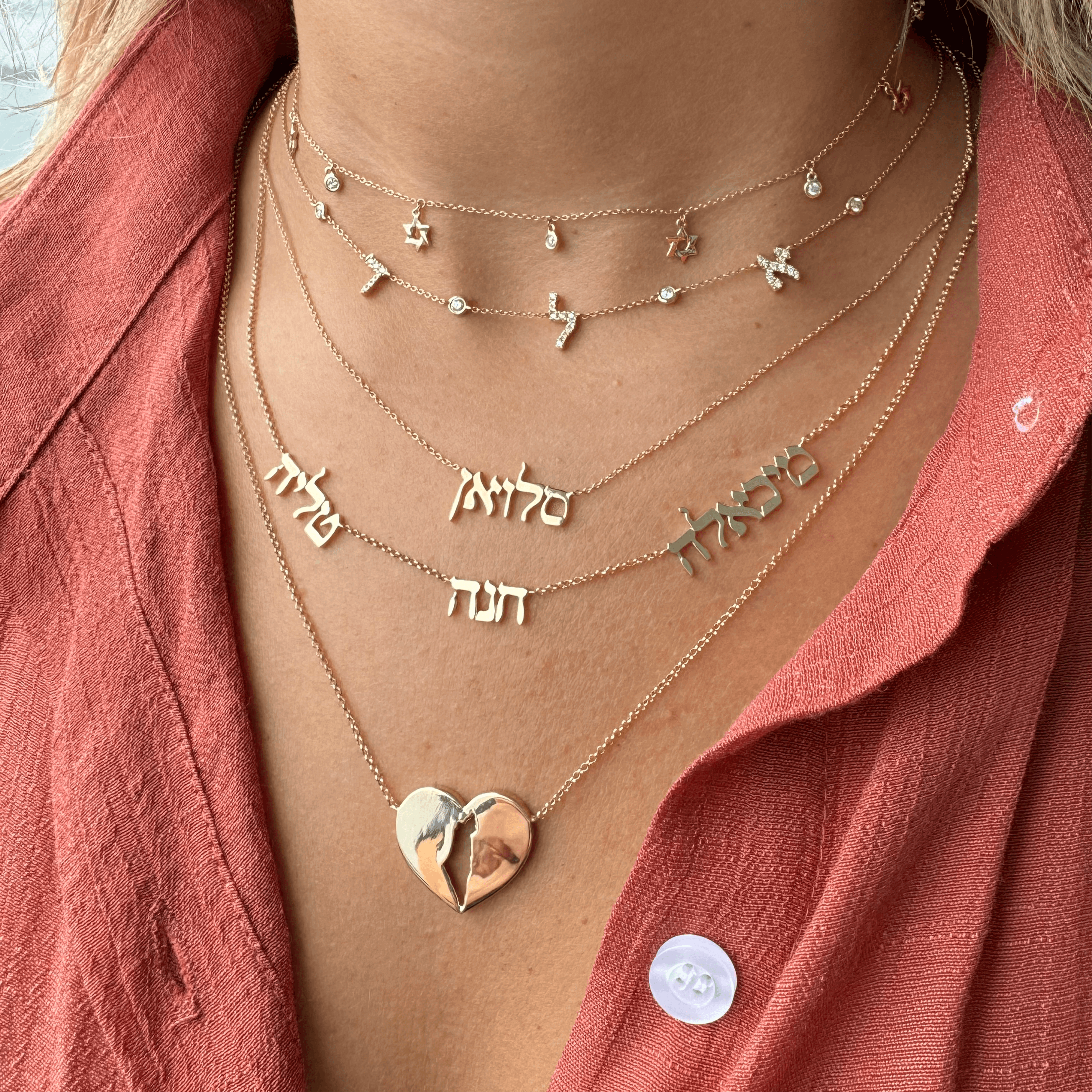 Woman wearing layered gold necklaces with personalized name pendants and a heart-shaped locket, styled with a coral shirt.