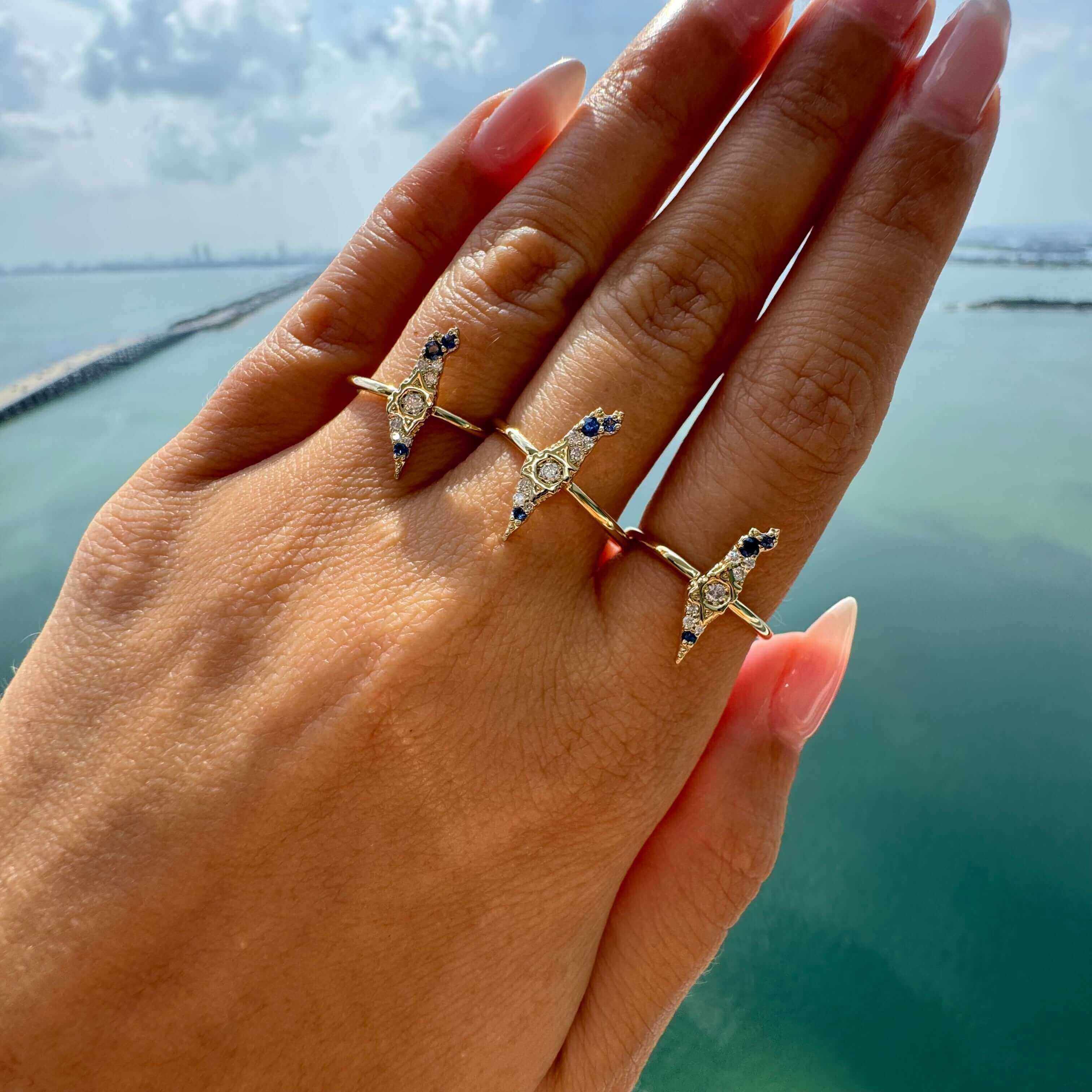 Elegant gold rings with intricate designs and gemstones shining on a hand near a scenic waterfront view.
