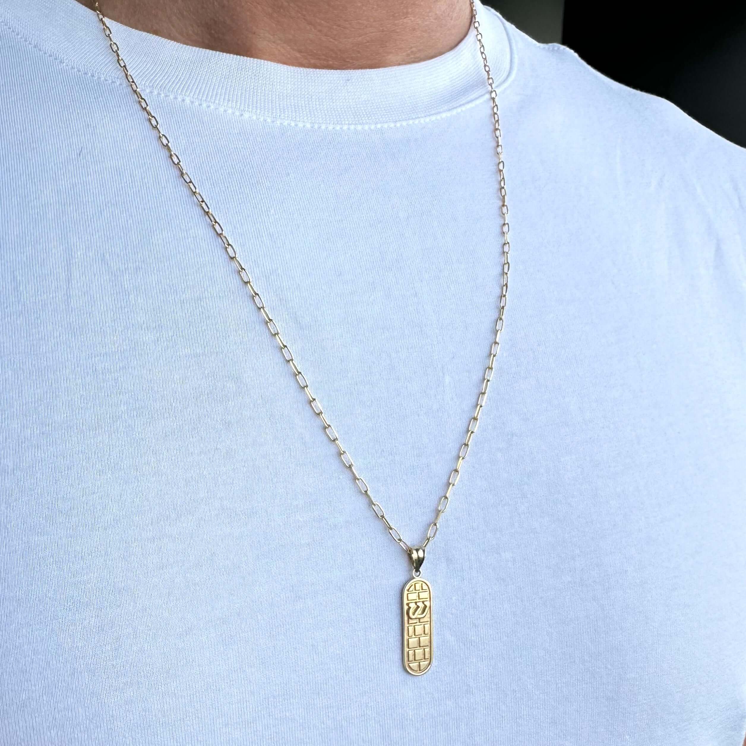 Close-up of a gold pendant necklace with a textured rectangular charm on a man's chest, showcasing elegant jewelry design.
