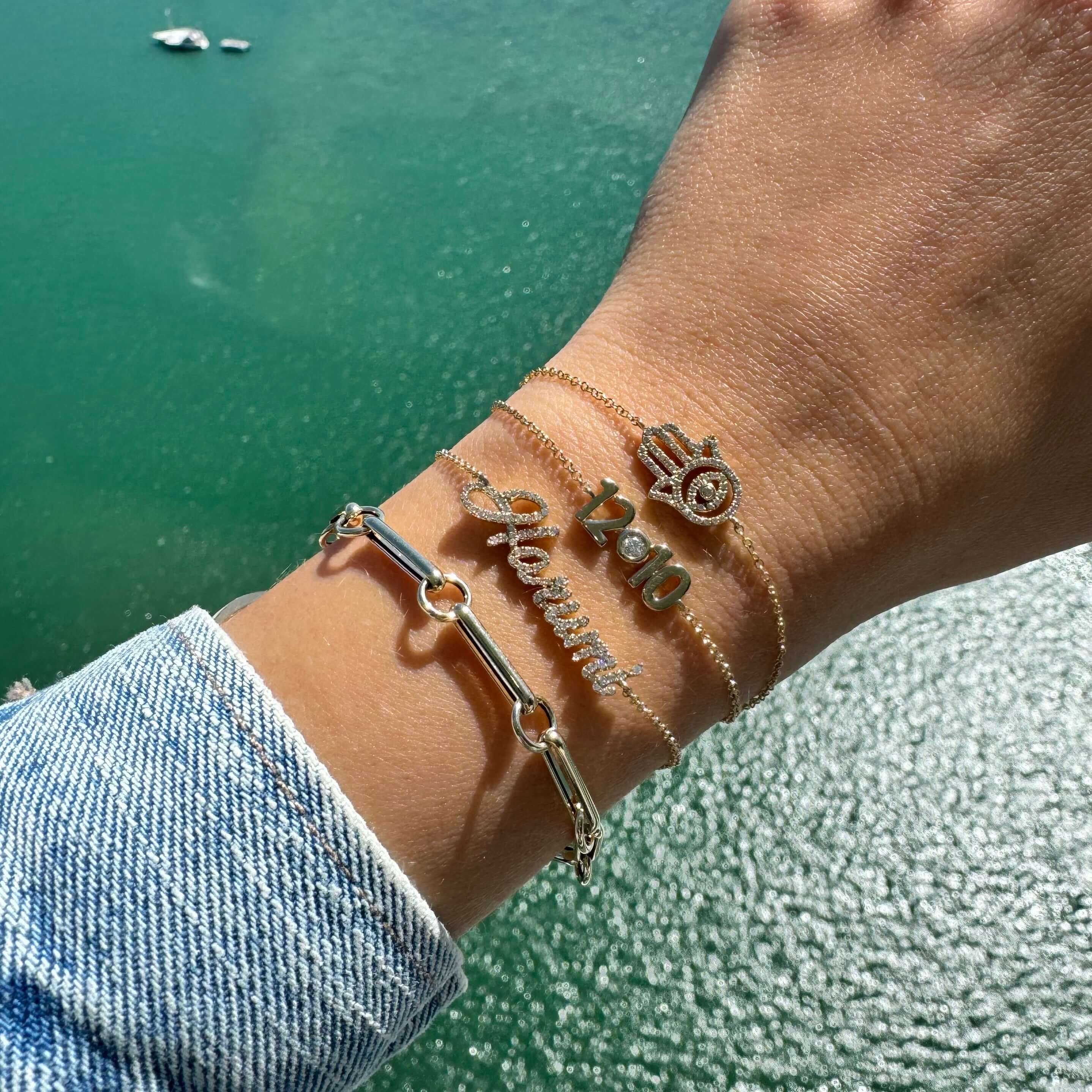 Fashionable stacked bracelets including 14K gold paper clip rolo link chain on a wrist beside a scenic water background.