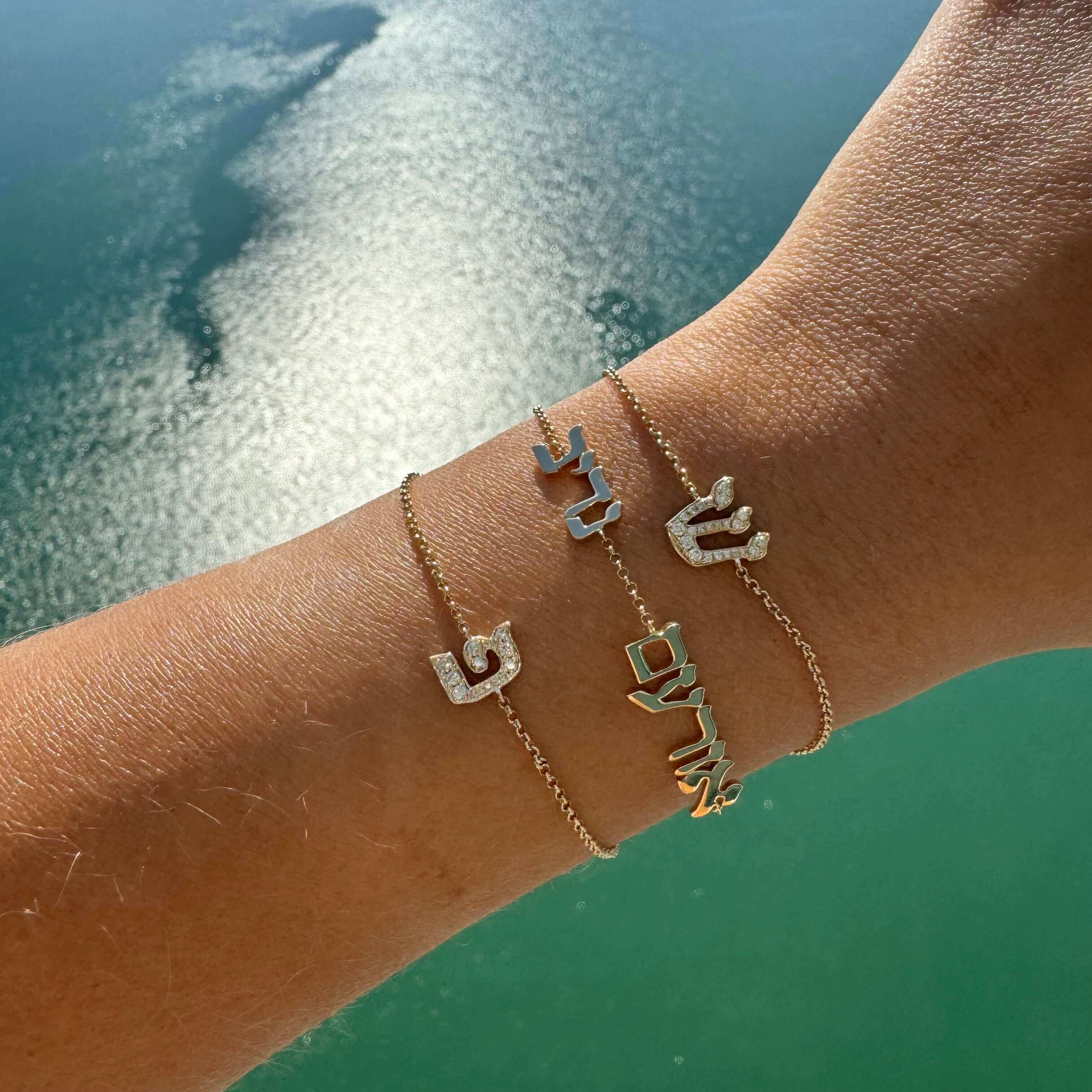 Stylish gold charm bracelets with initials, worn on wrist, showcased against a serene ocean background, perfect for personalized jewelry lovers.