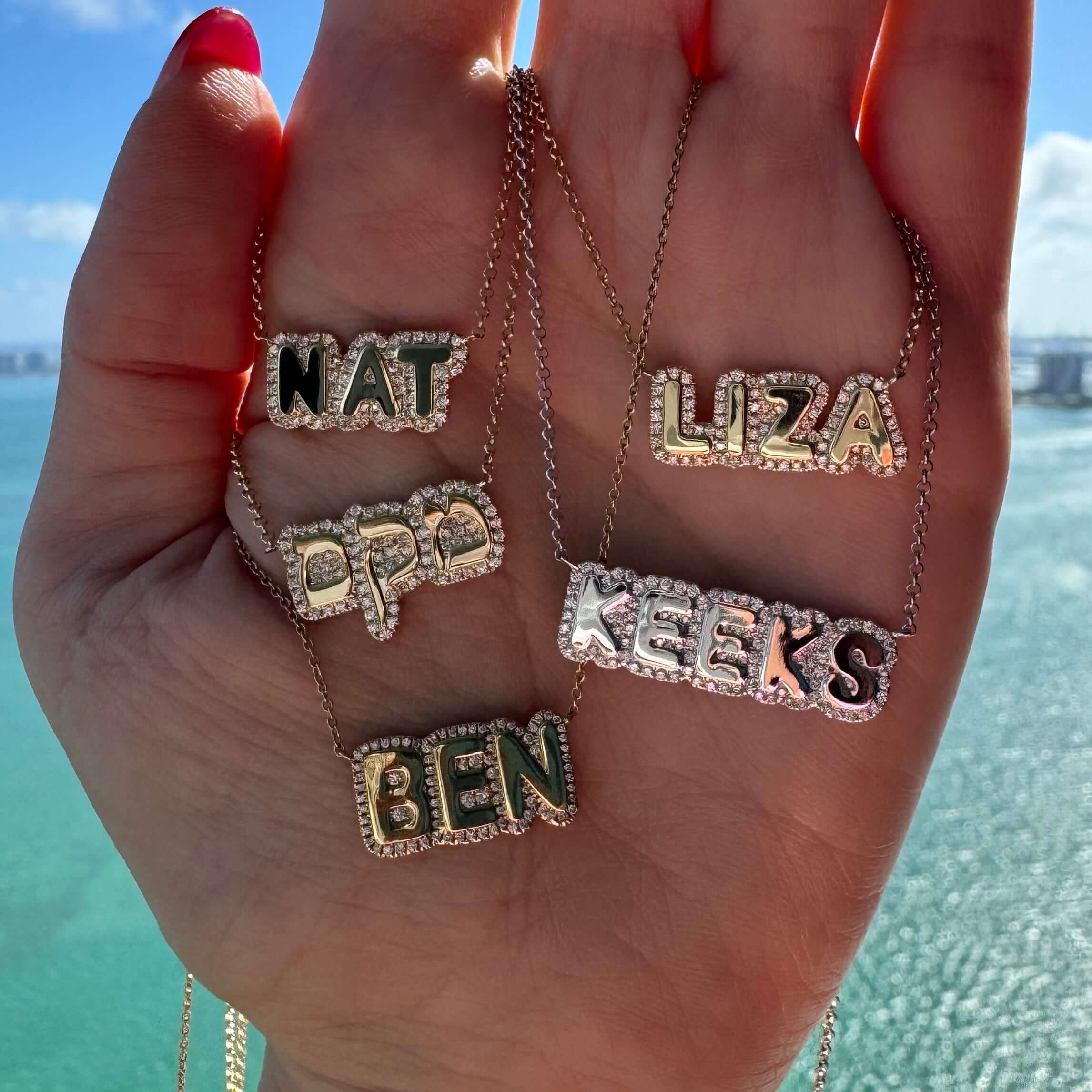 Personalized nameplate necklaces in 14K gold, featuring names NAT, LIZA, BEN, and KEKS against a scenic ocean backdrop.