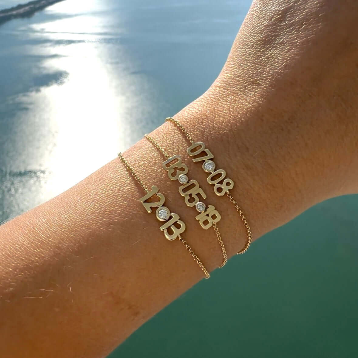 Gold bracelets with custom date designs and diamond accents, showcased on a wrist near a serene body of water, from our jewelry collection.