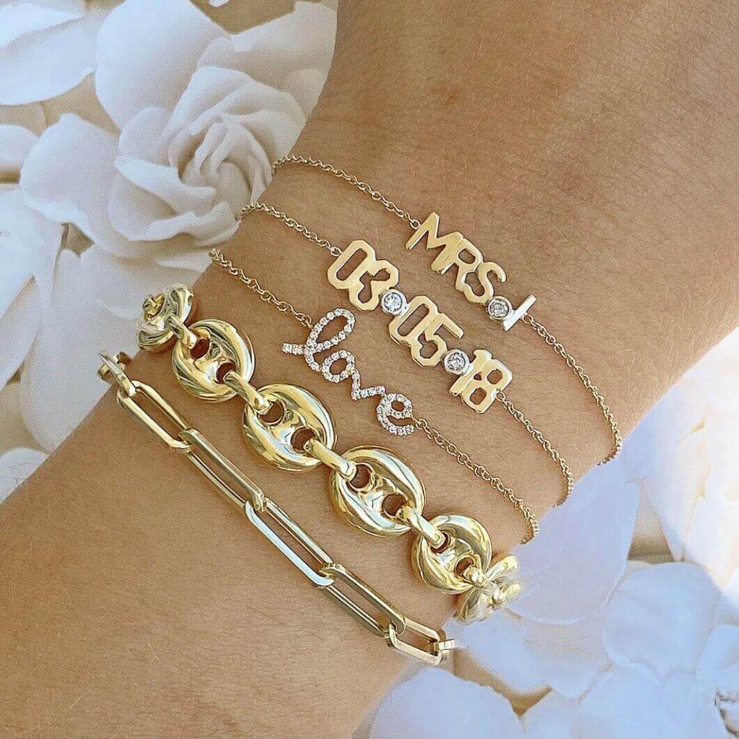 Elegant gold and diamond bracelets showcasing vibrant lettering designs and bold chains, displayed on a wrist with white floral background.