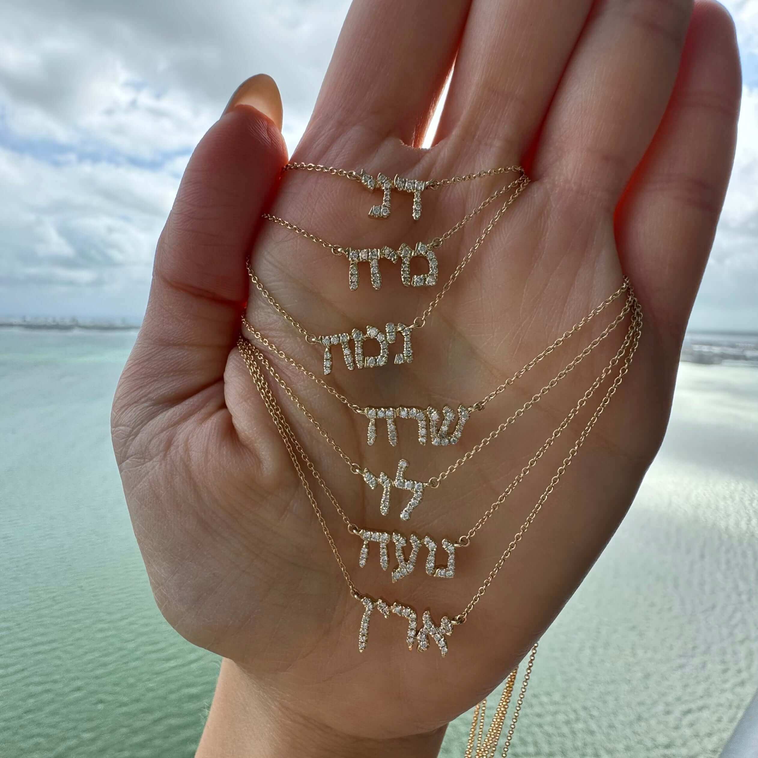Hand holding multiple Hebrew name necklaces over water background, showcasing elegant and personalized jewelry from our brand