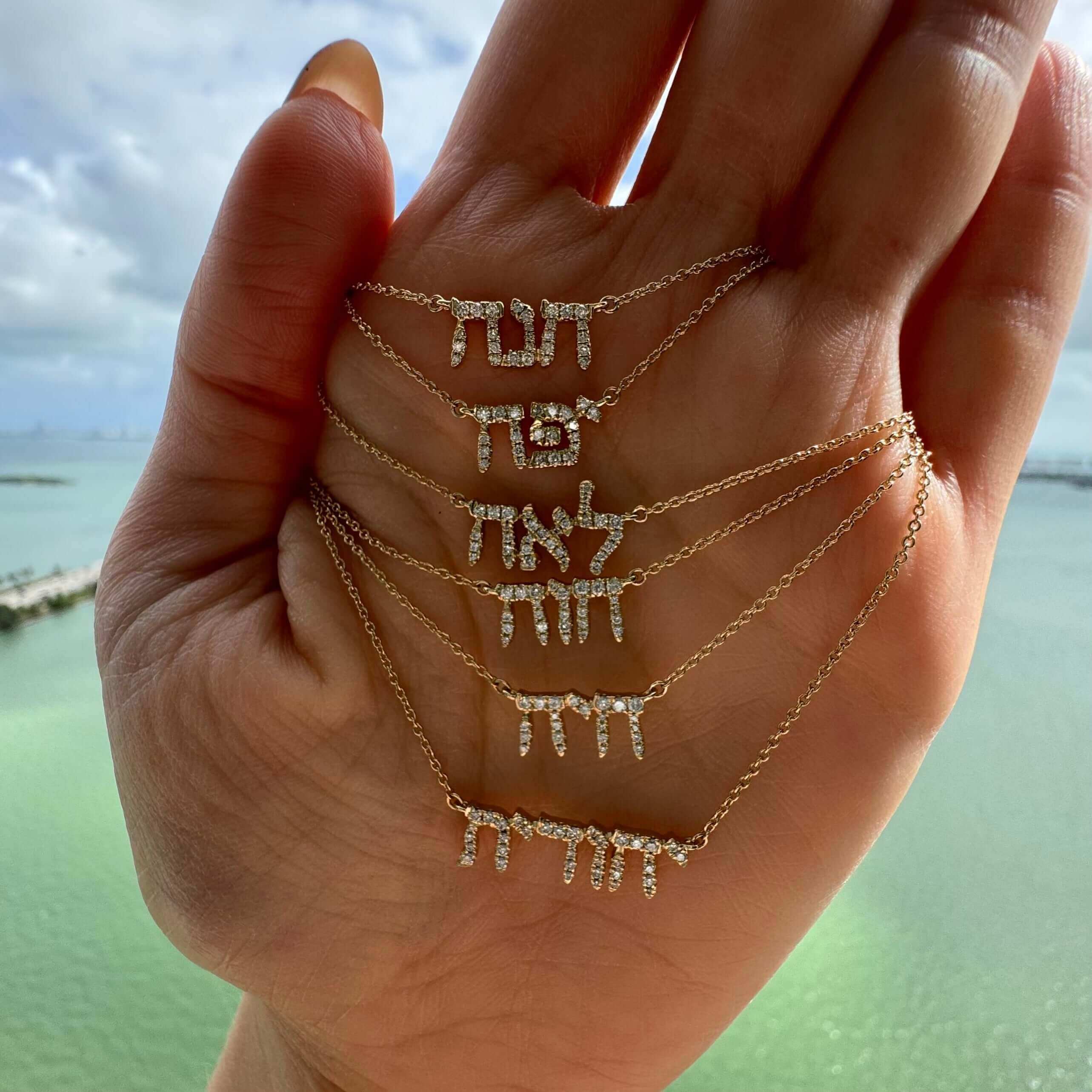 Hand displaying elegant Hebrew letter necklaces by the sea.