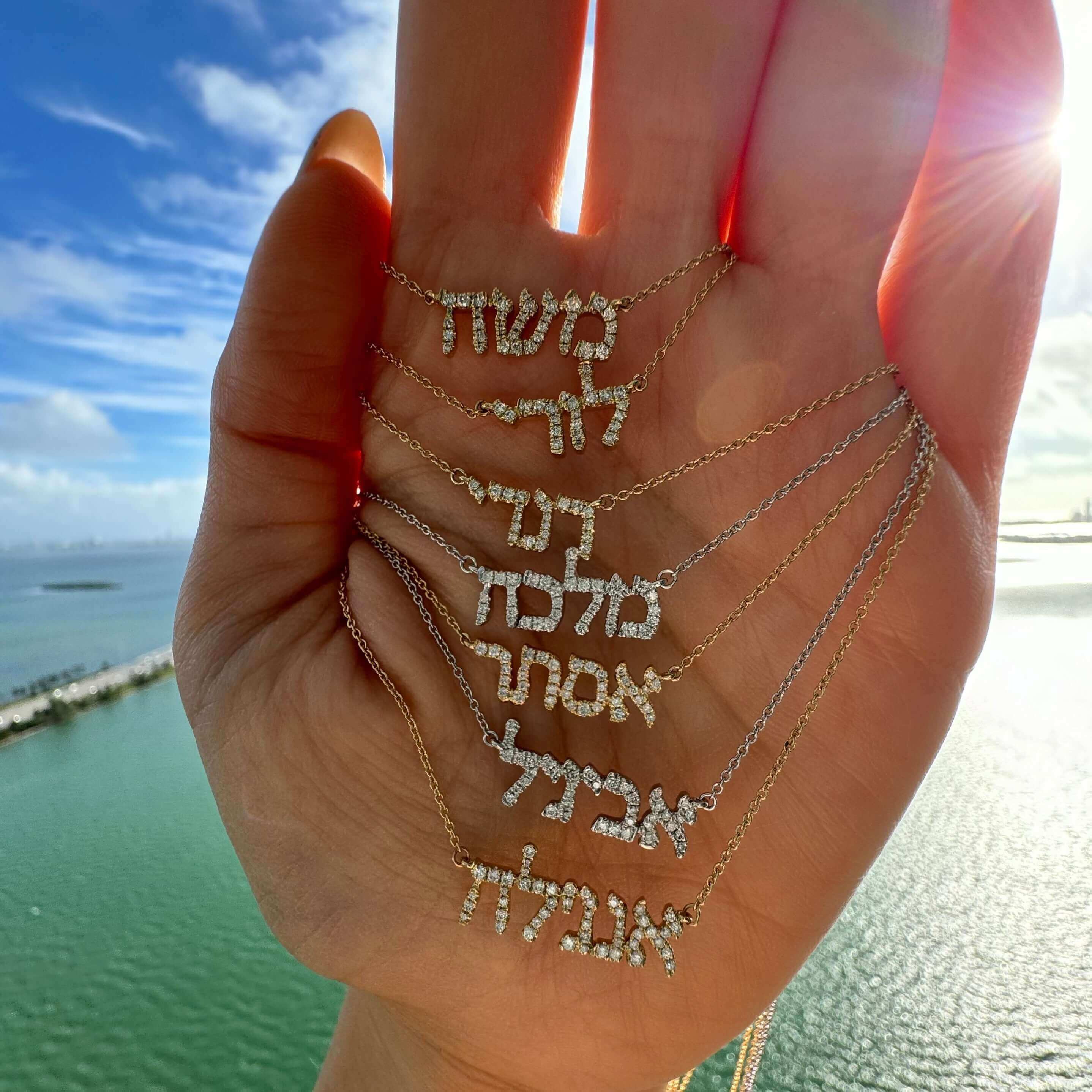 Personalized Hebrew Diamond Nameplate Necklace displayed in hand against a scenic water background, Izakov Fine Jewelry.