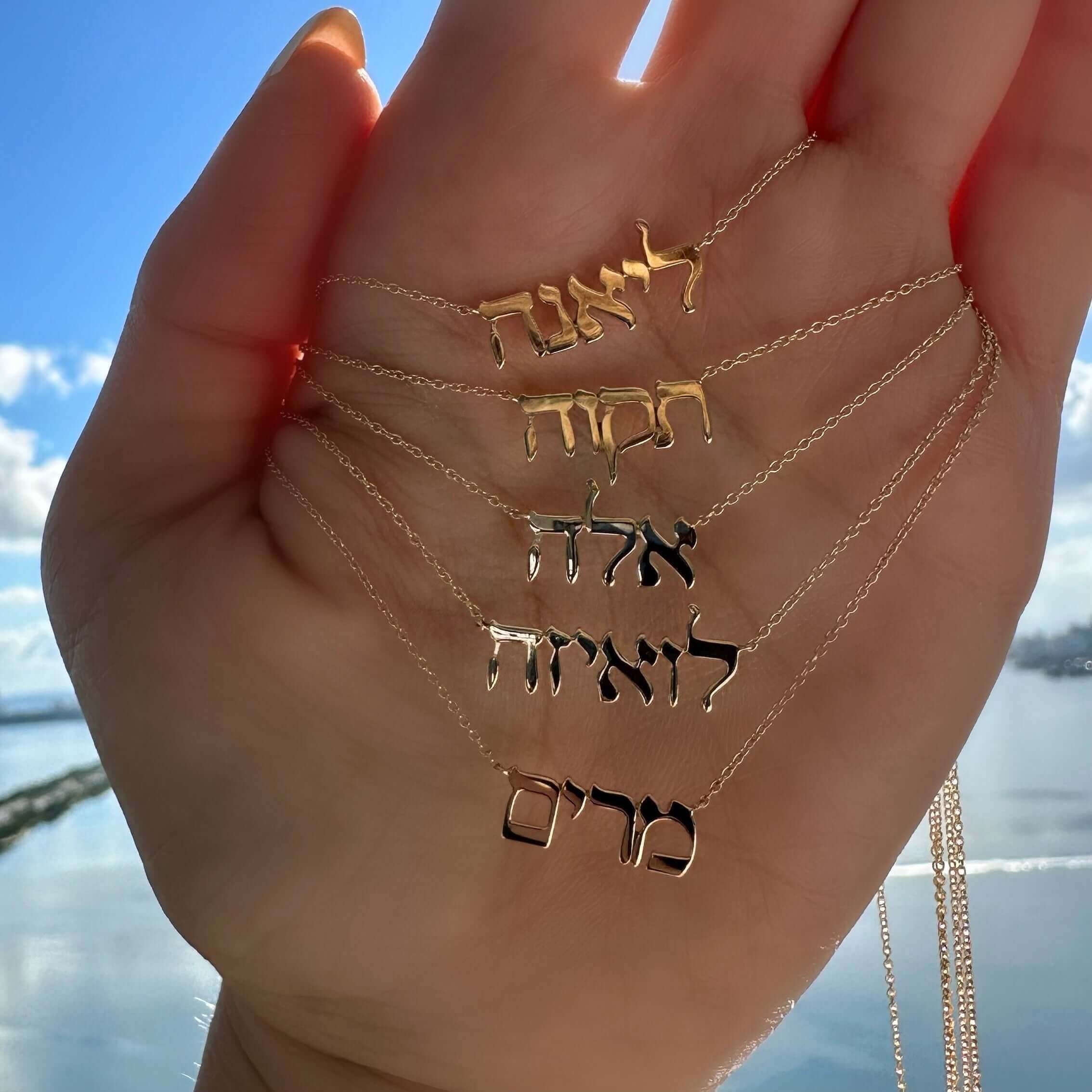 Gold and silver personalized name necklaces from our jewelry brand displayed on a hand with a scenic water background.