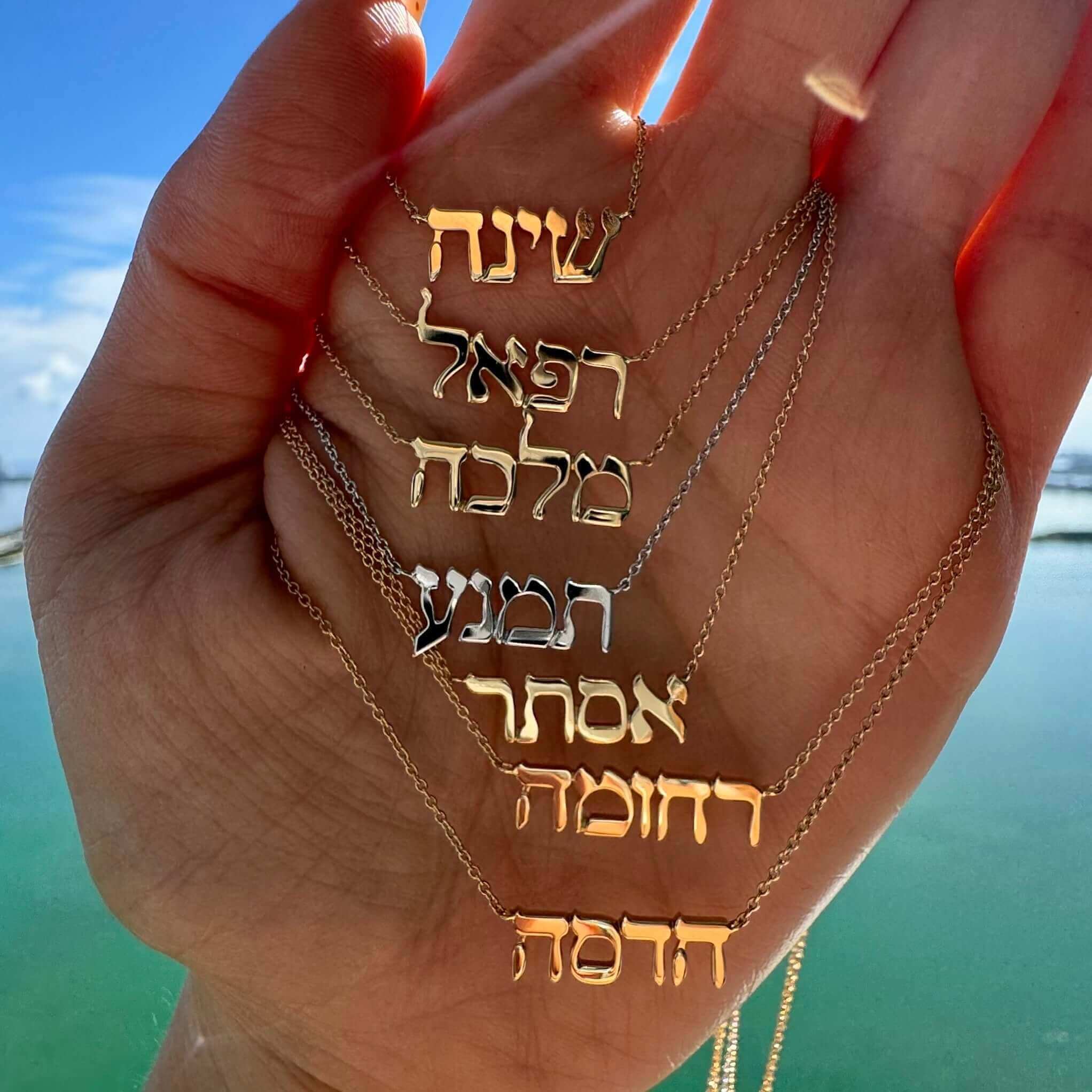 Hand holding gold and silver name necklaces from our jewelry brand, with a scenic sky background.