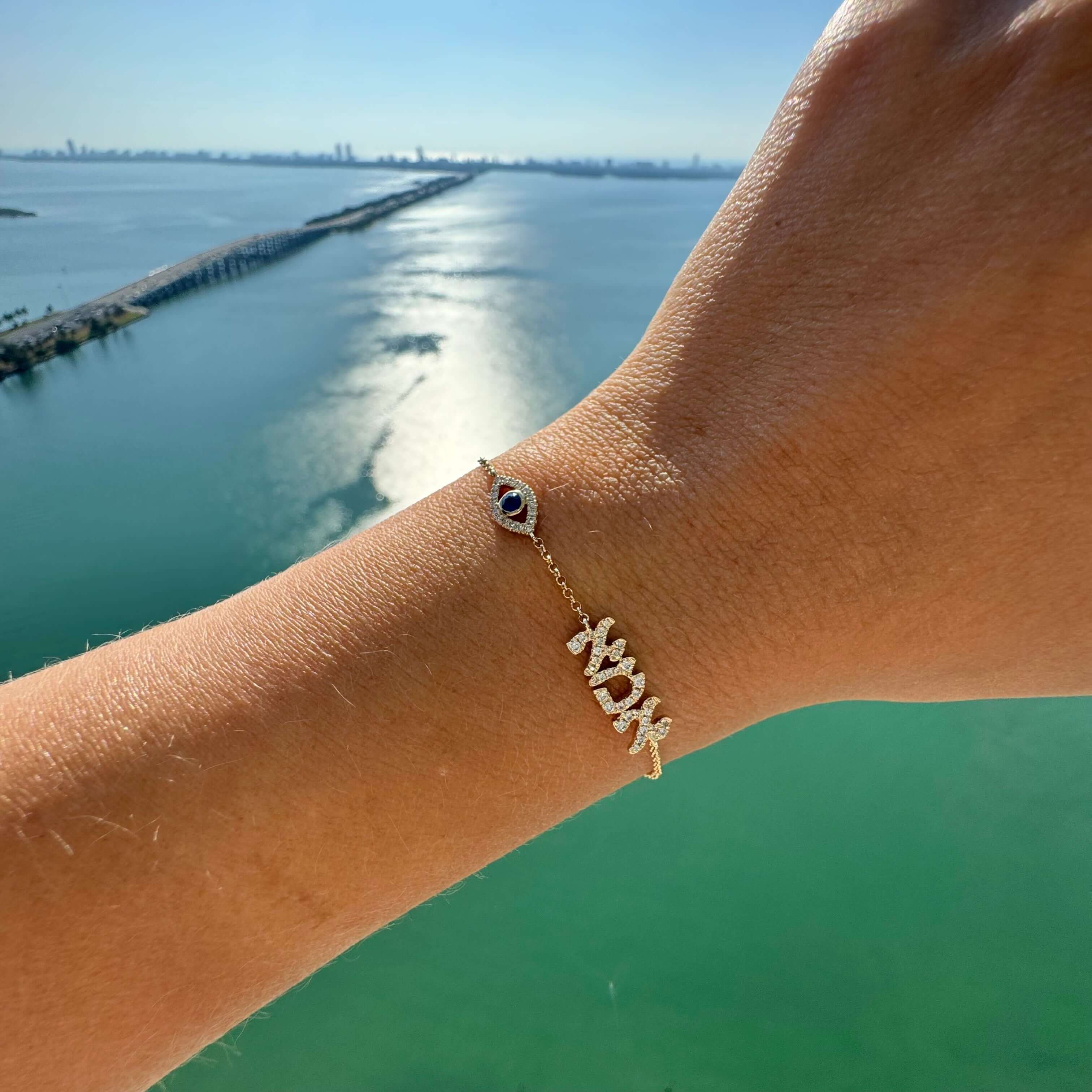 Personalized Motif Hebrew Diamond Nameplate Bracelet with sapphire evil eye on a wrist against a scenic ocean background.
