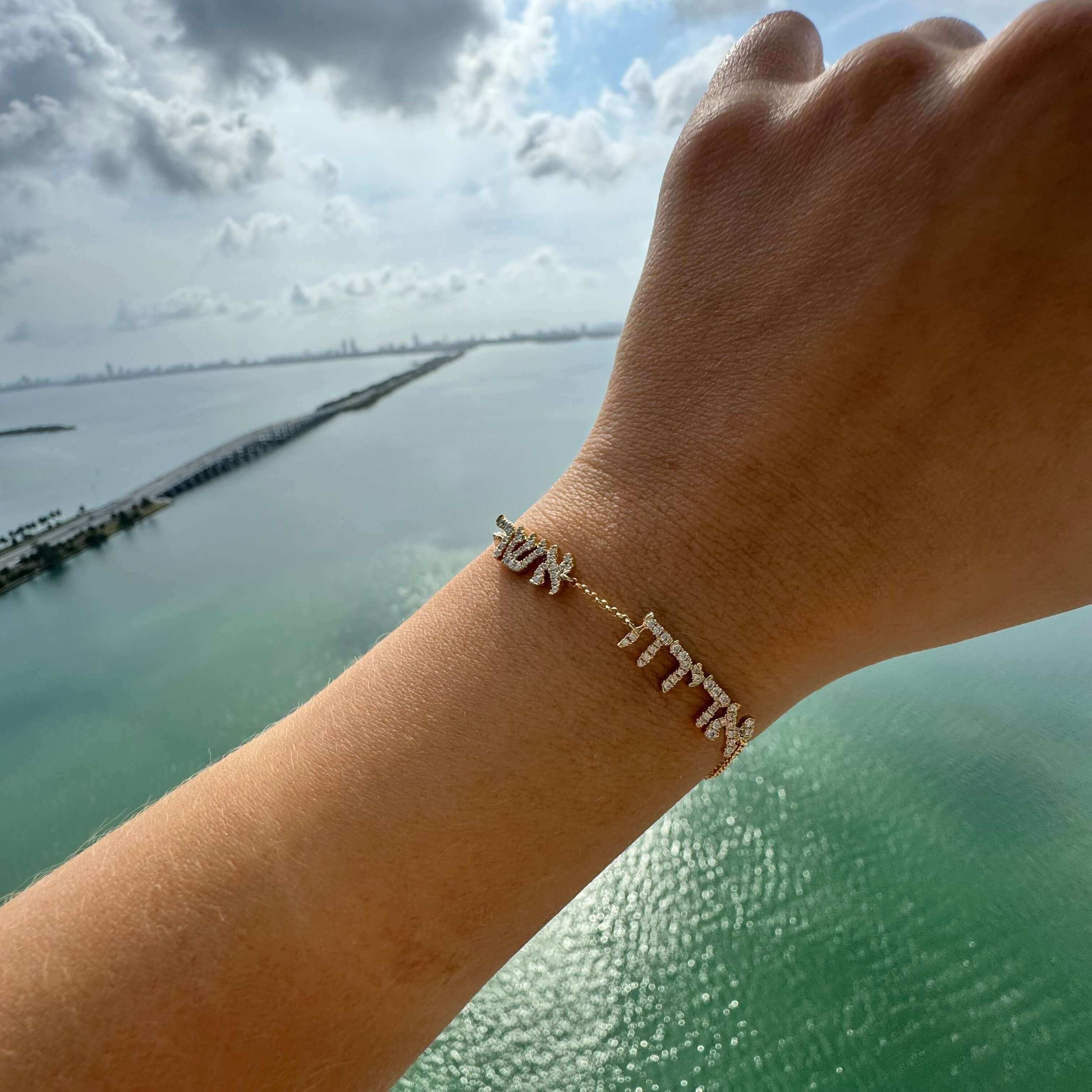 Elegant gold bracelet with diamond-studded charms spelling "SUN" and "FUN" showcased above a scenic ocean view.