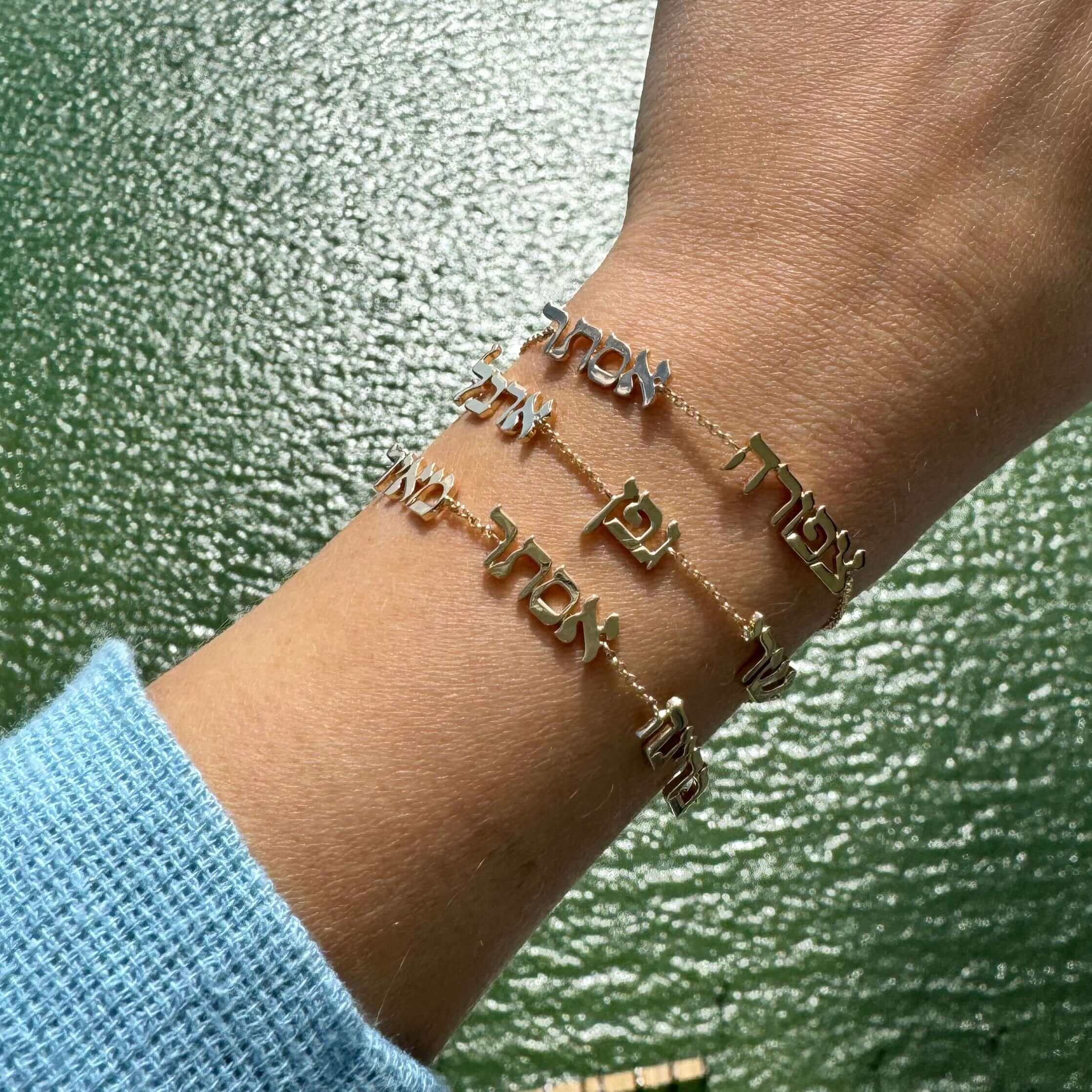 Woman's wrist adorned with elegant gold script bracelets from our jewelry brand, sparkling against a serene green water background.