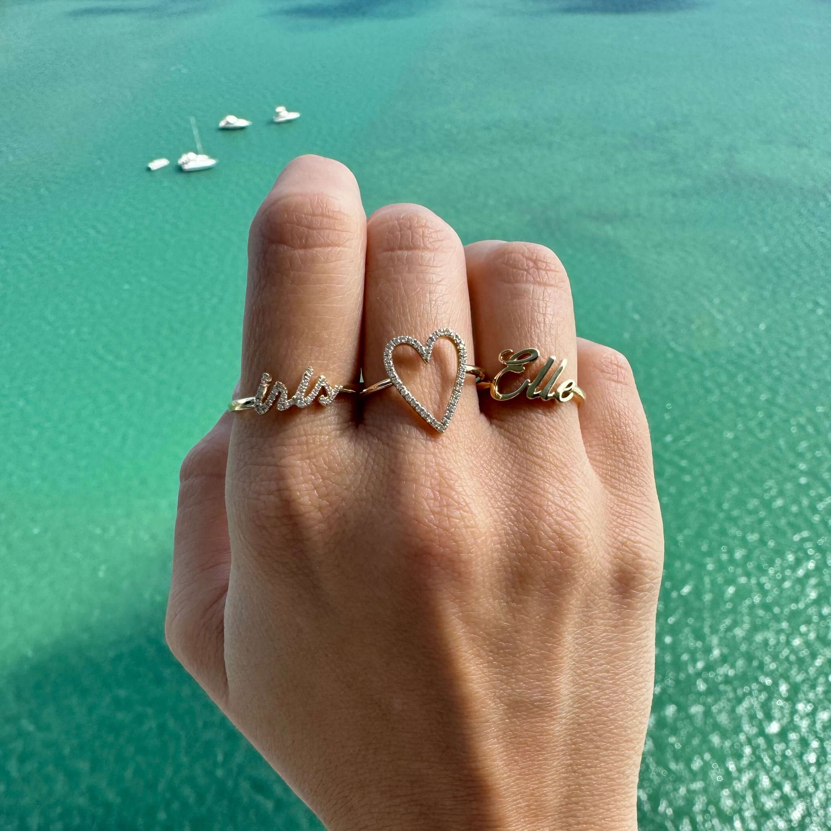 14K Gold personalized script nameplate rings with diamonds on hand against turquoise water background.