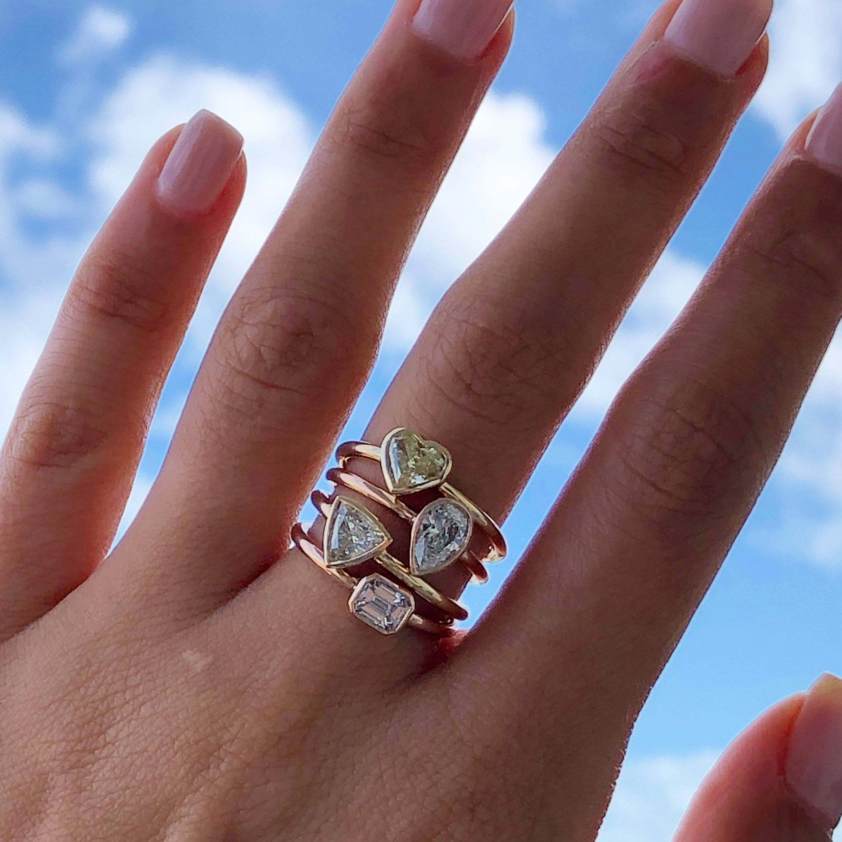 Hand wearing four gold rings with various diamond shapes from our luxury jewelry collection against a blue sky background.