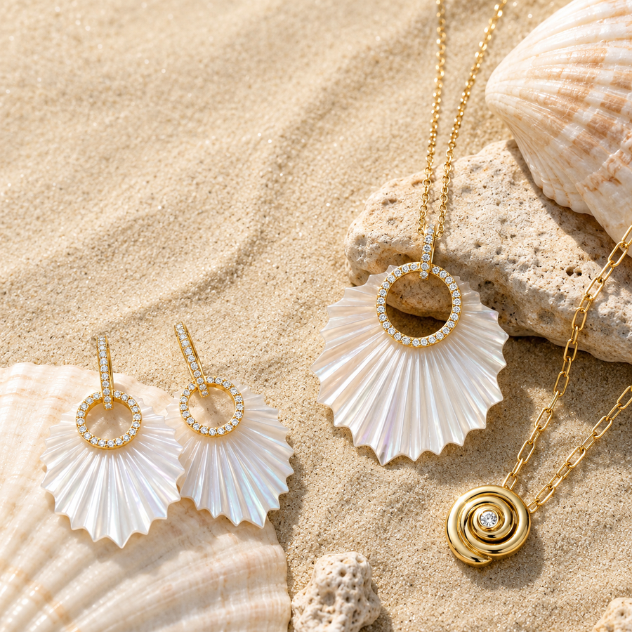 Gold jewelry set with white iridescent shell-inspired pendants and drop earrings, arranged on sand with seashells and rocks. The set includes a fan-shaped shell pendant necklace, matching drop earrings with ring-shaped settings, and a smaller gold spiral pendant.