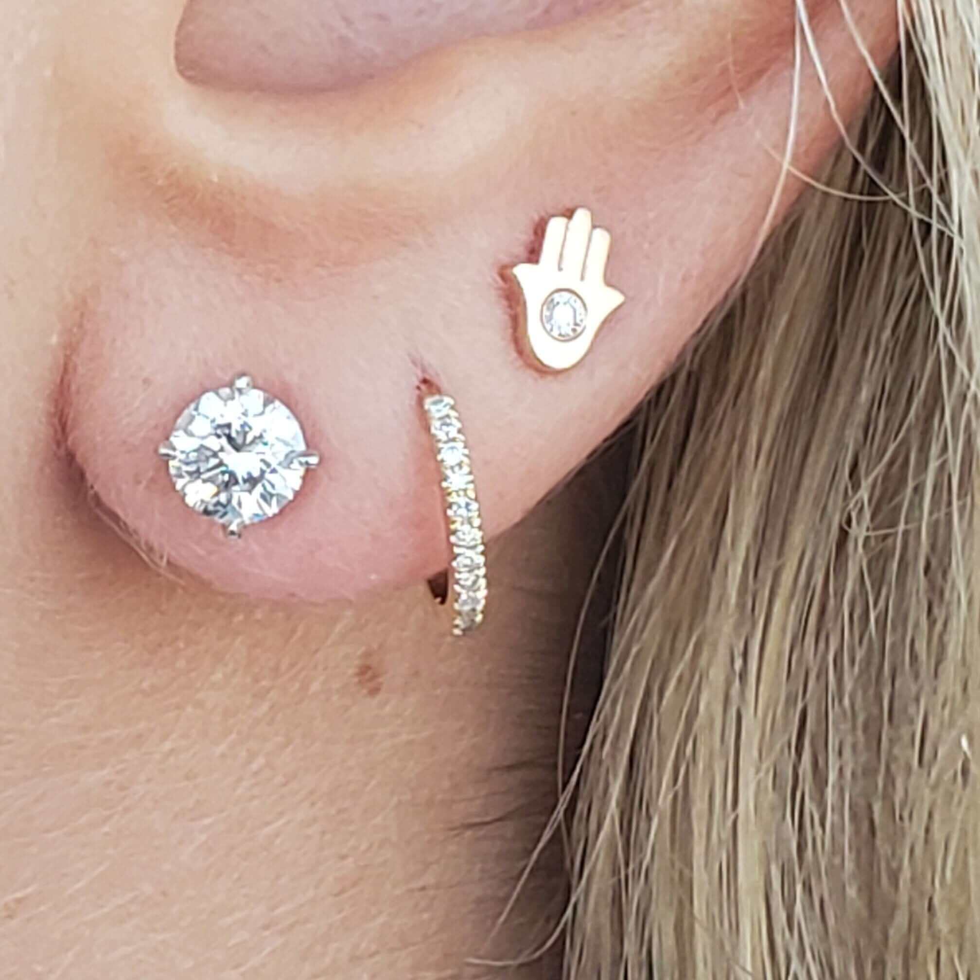 Close-up of triple earring set featuring a sparkling diamond stud, a gold hoop with small diamonds, and a gold Hamsa hand with a diamond detail.