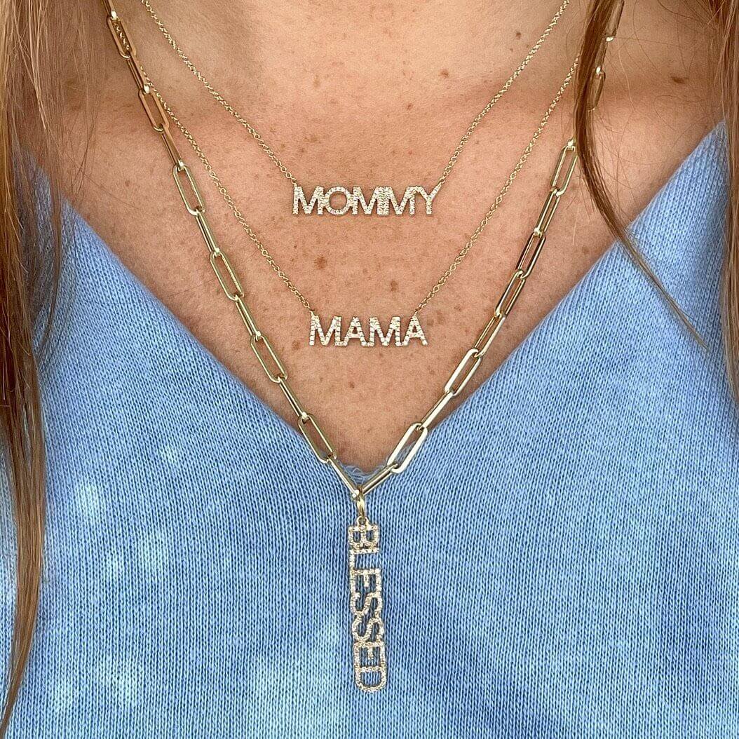Layered gold necklaces with "MOMMY," "MAMA," and "BLESSED" pendants on blue blouse, representing our elegant and personalized jewelry collection.