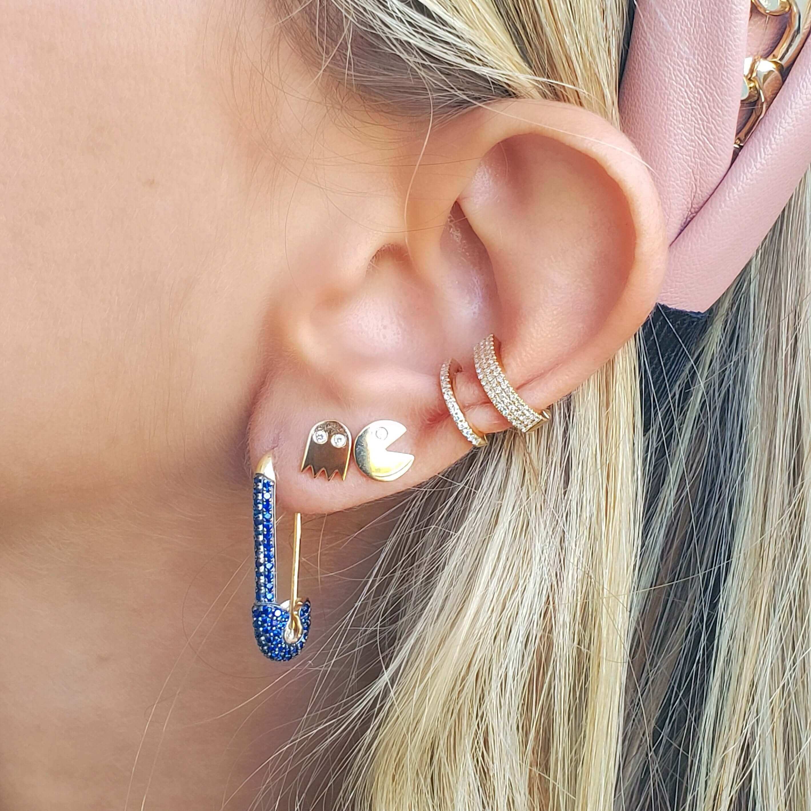 Woman wearing a set of trendy earrings including gold hoop, Pac-Man design, and crystal-encrusted safety pin, showcasing unique jewelry style.