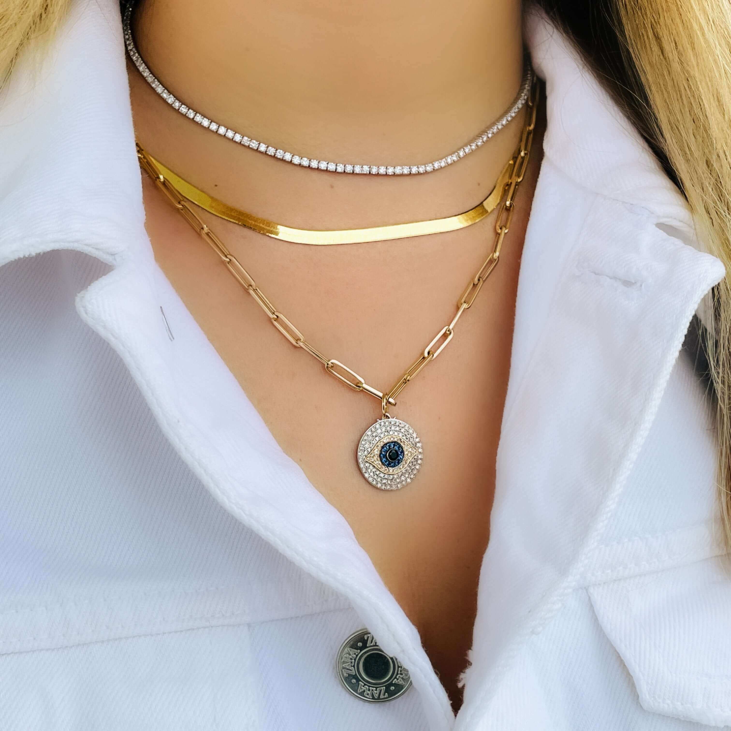 Woman wearing layered gold and diamond necklaces with a white shirt, featuring a blue and white gemstone pendant.