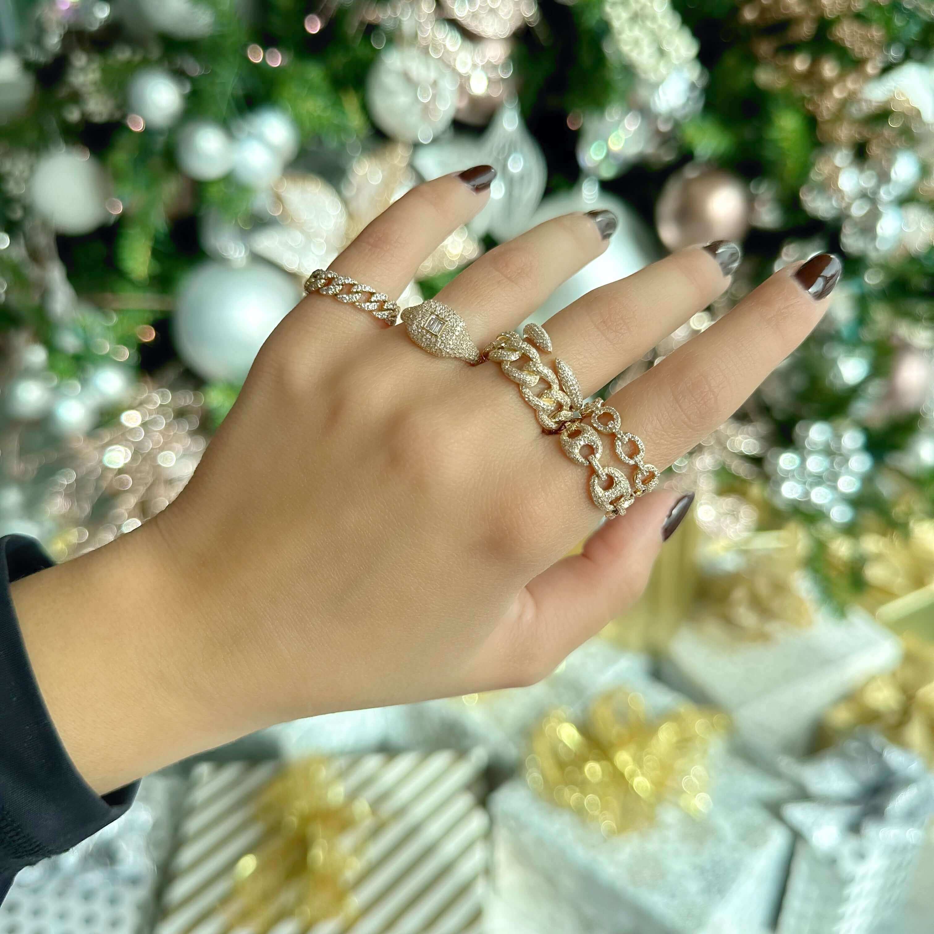 Hand adorned with elegant gold rings featuring intricate designs, surrounded by a festive holiday backdrop with wrapped gifts and decorations.