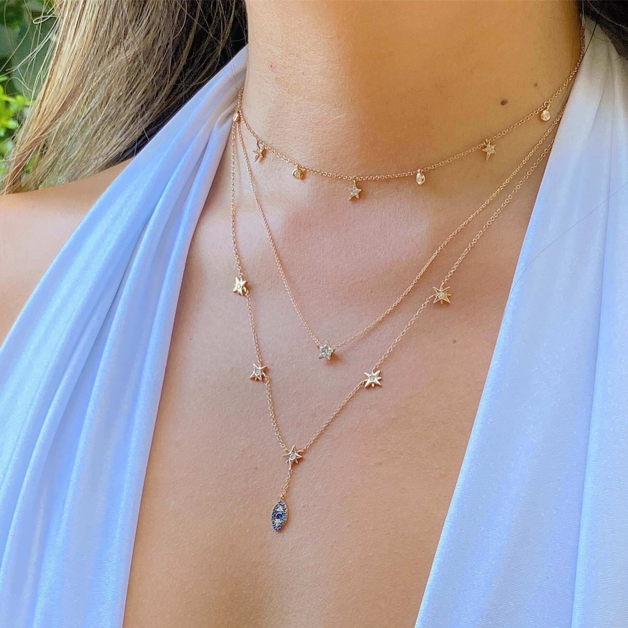 Woman wearing layered gold necklaces with star and eye pendants, styled with a white halter top, showcasing elegant jewelry design.