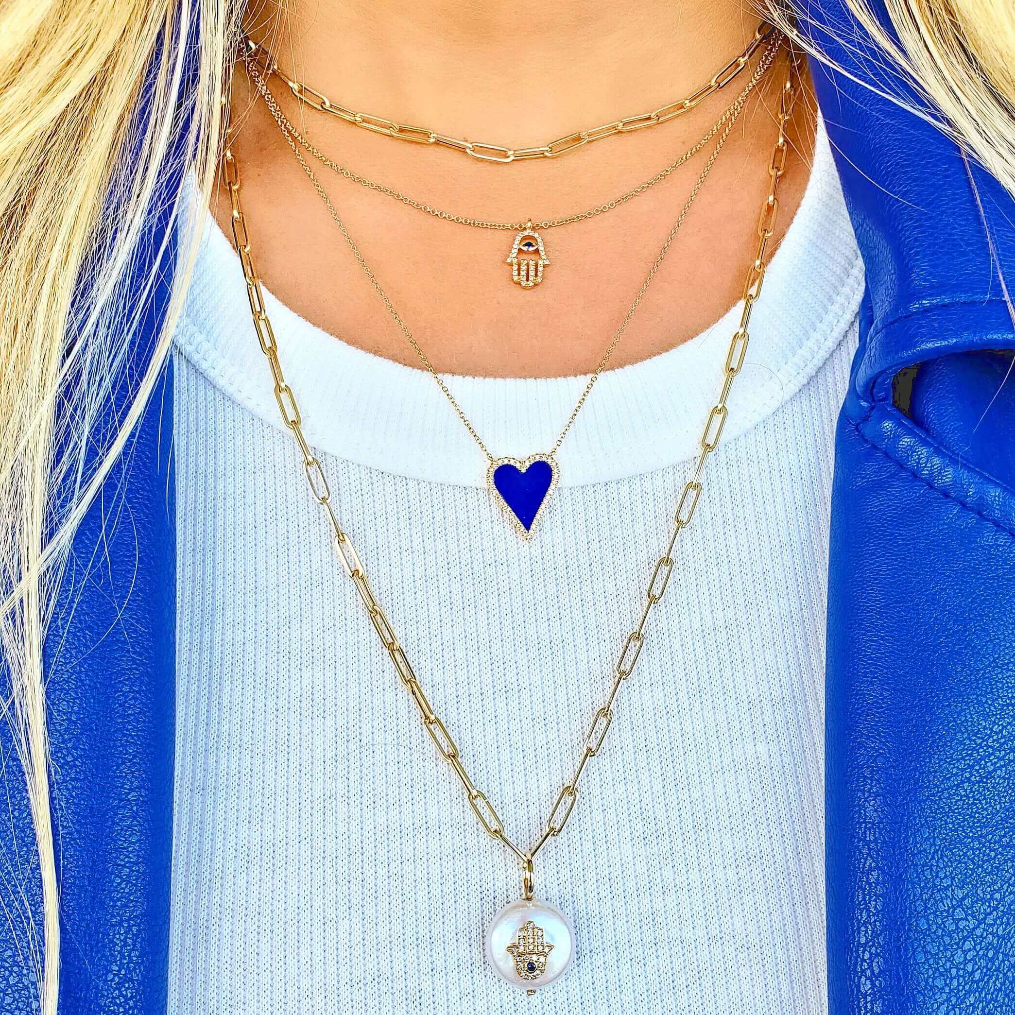 Layered gold necklaces with heart and hamsa pendants styled on a woman wearing a blue jacket and white top, showcasing our luxury jewelry brand.