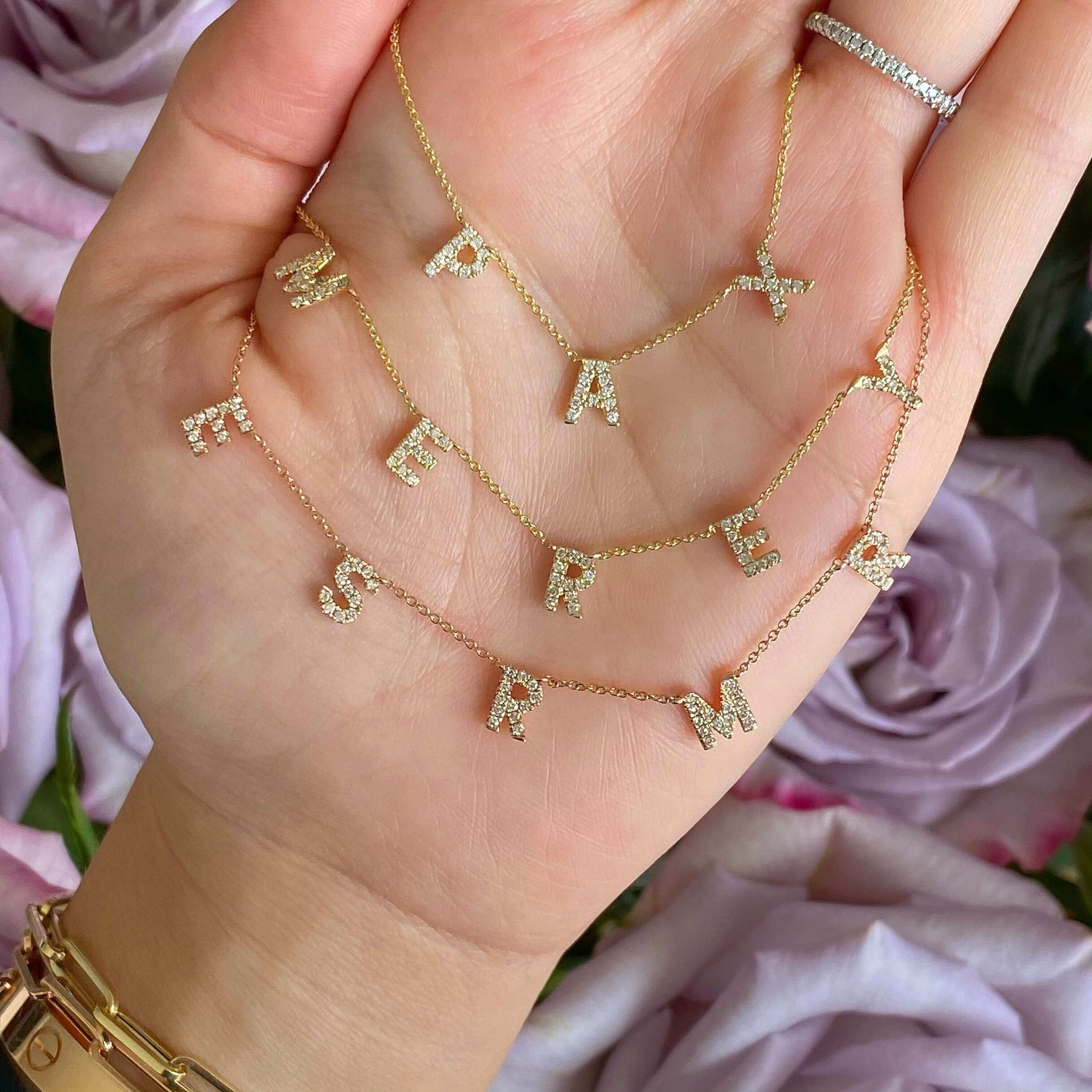 Woman holding gold letter necklaces with diamond accents from our jewelry collection, surrounded by soft pink roses.