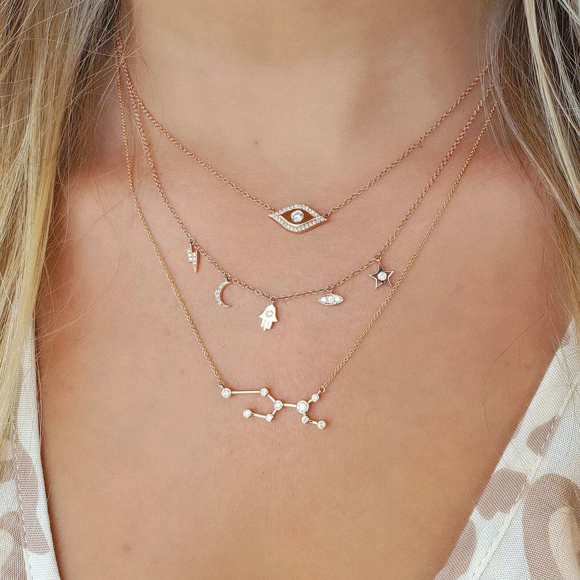 Woman wearing layered gold chain necklaces featuring various charms, including a constellation, crescent moon, star, and protective eye symbol.