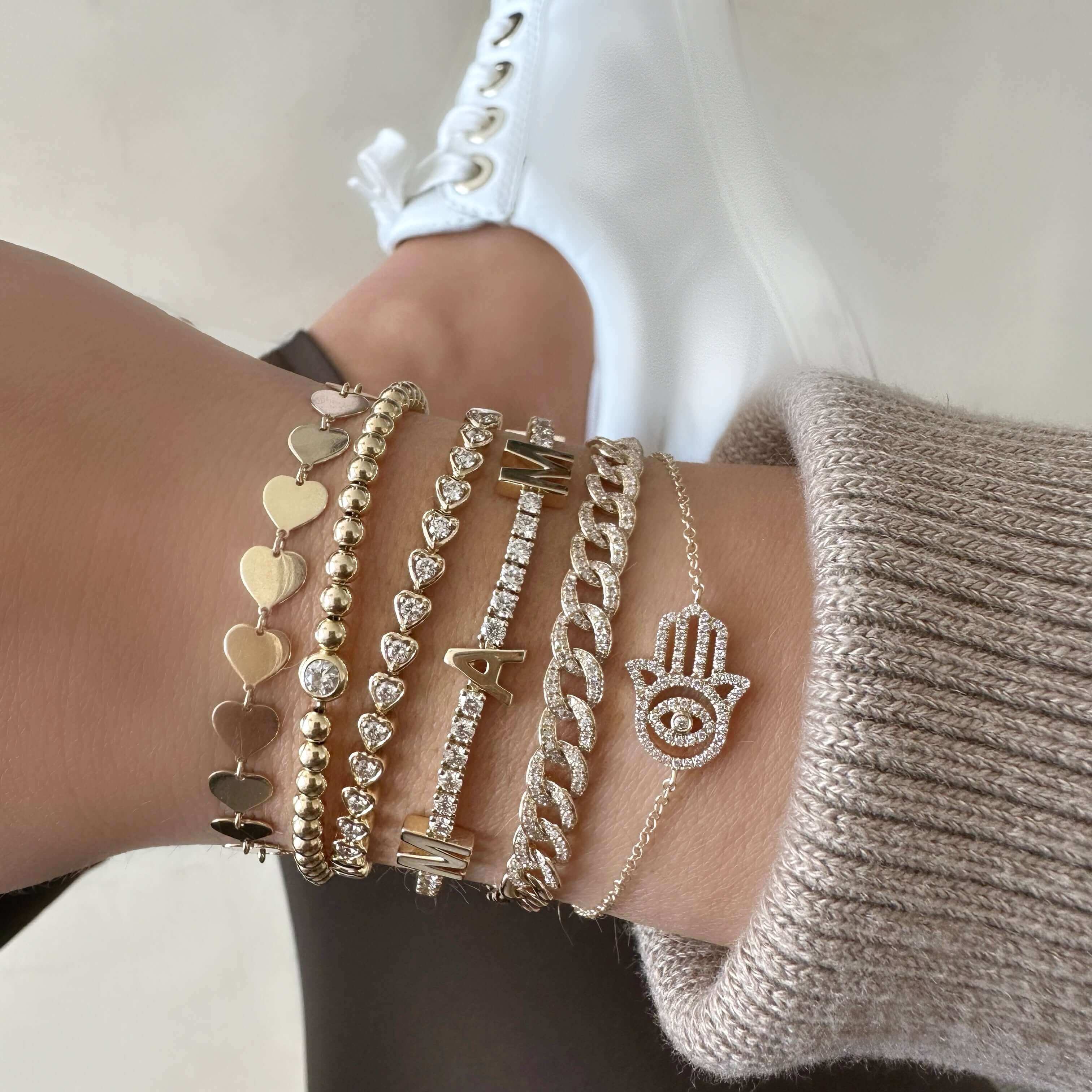 Elegant gold and diamond jewelry bracelets stacked on arm, featuring a hamsa hand charm, complementing casual fashion with white sneakers.