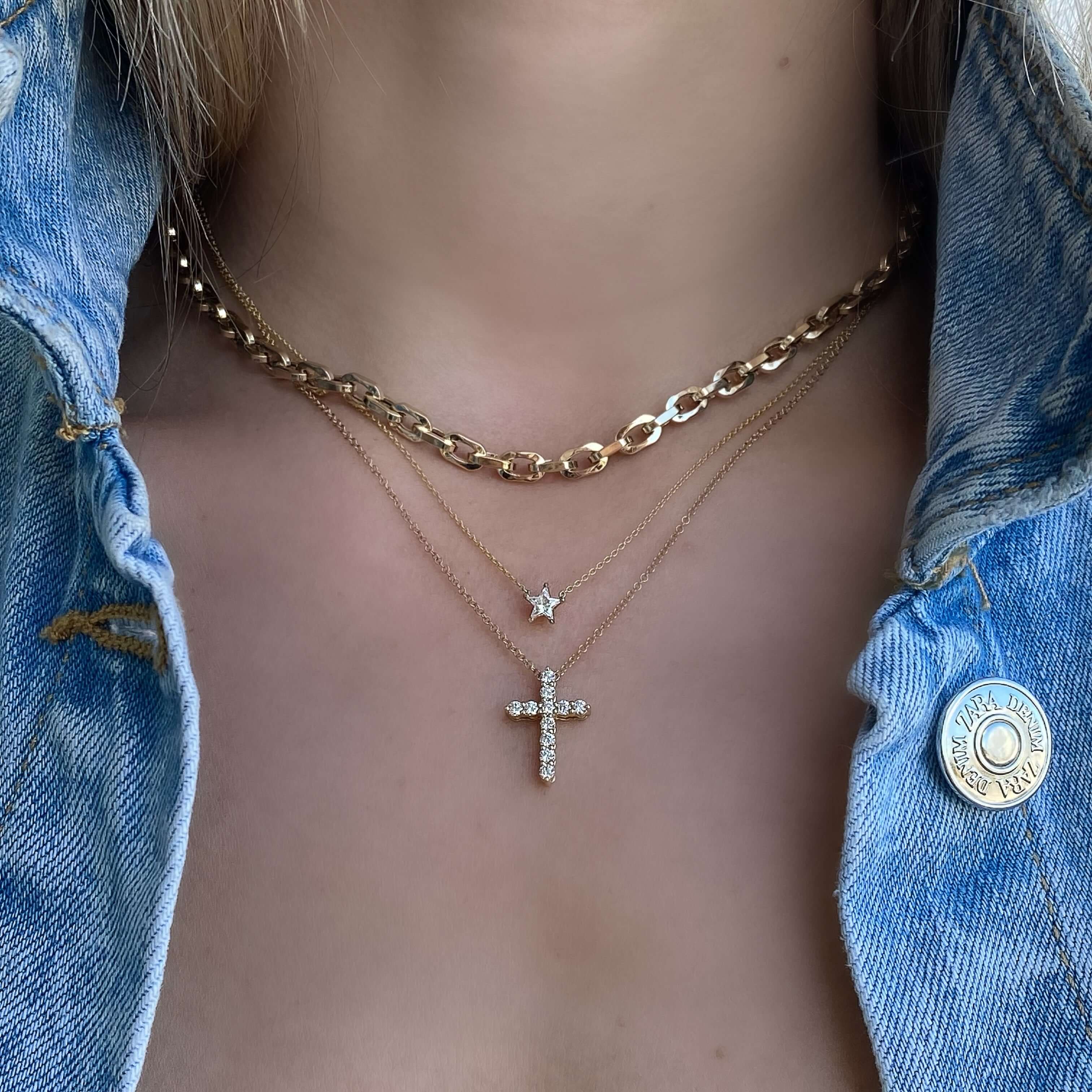 Woman wearing layered gold necklaces with cross pendant and star charm, styled over denim jacket.