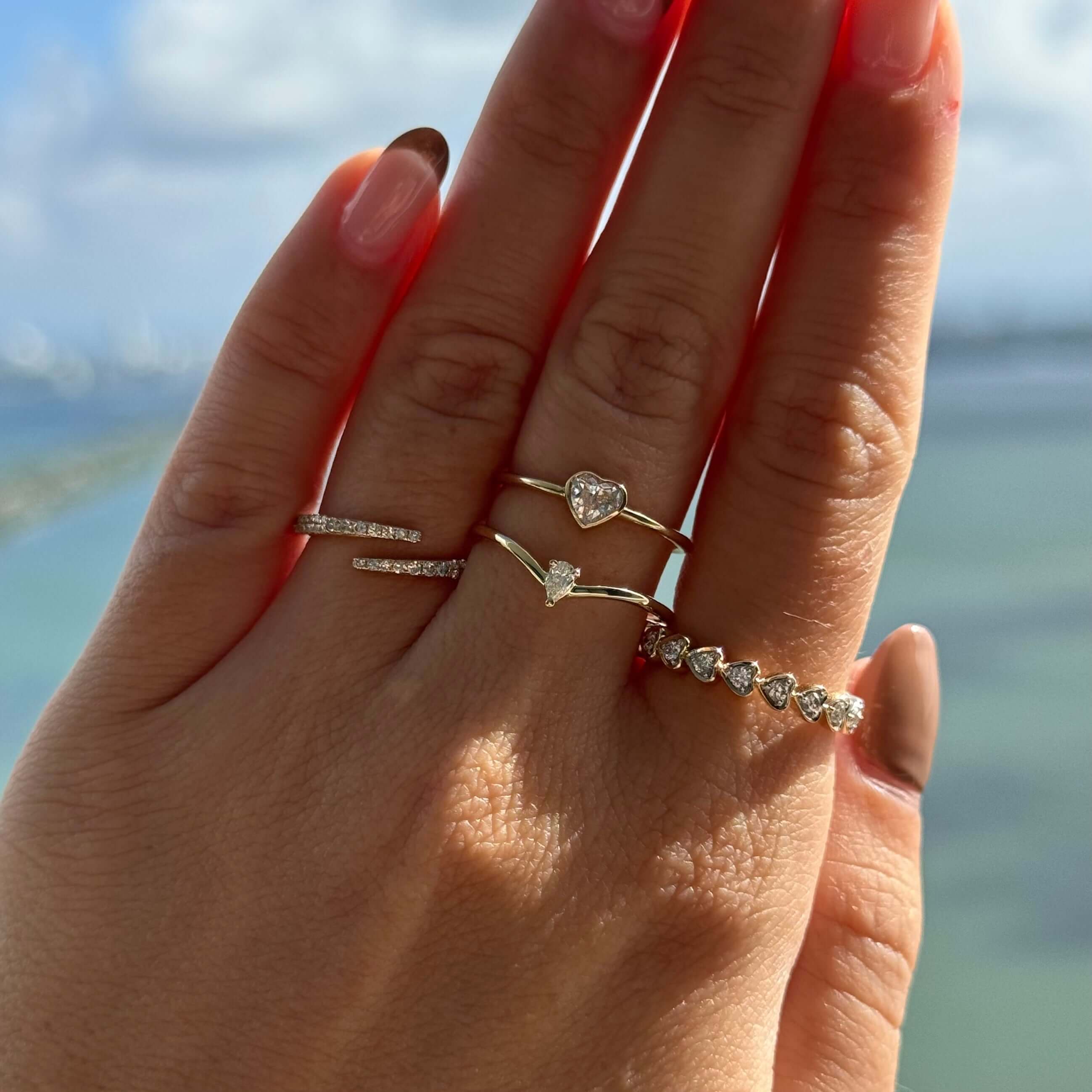 Hand wearing multiple delicate rings, including a heart-shaped diamond and curved gold designs, against a scenic background.