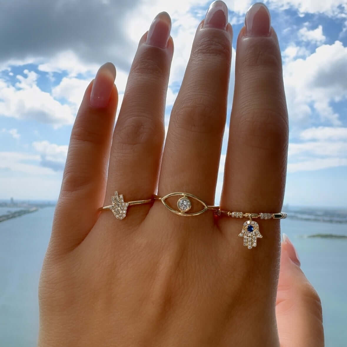 Woman's hand wearing three intricate Hamsa and Evil Eye gold rings with diamond accents, showcasing a stylish jewelry collection against a blue sky.