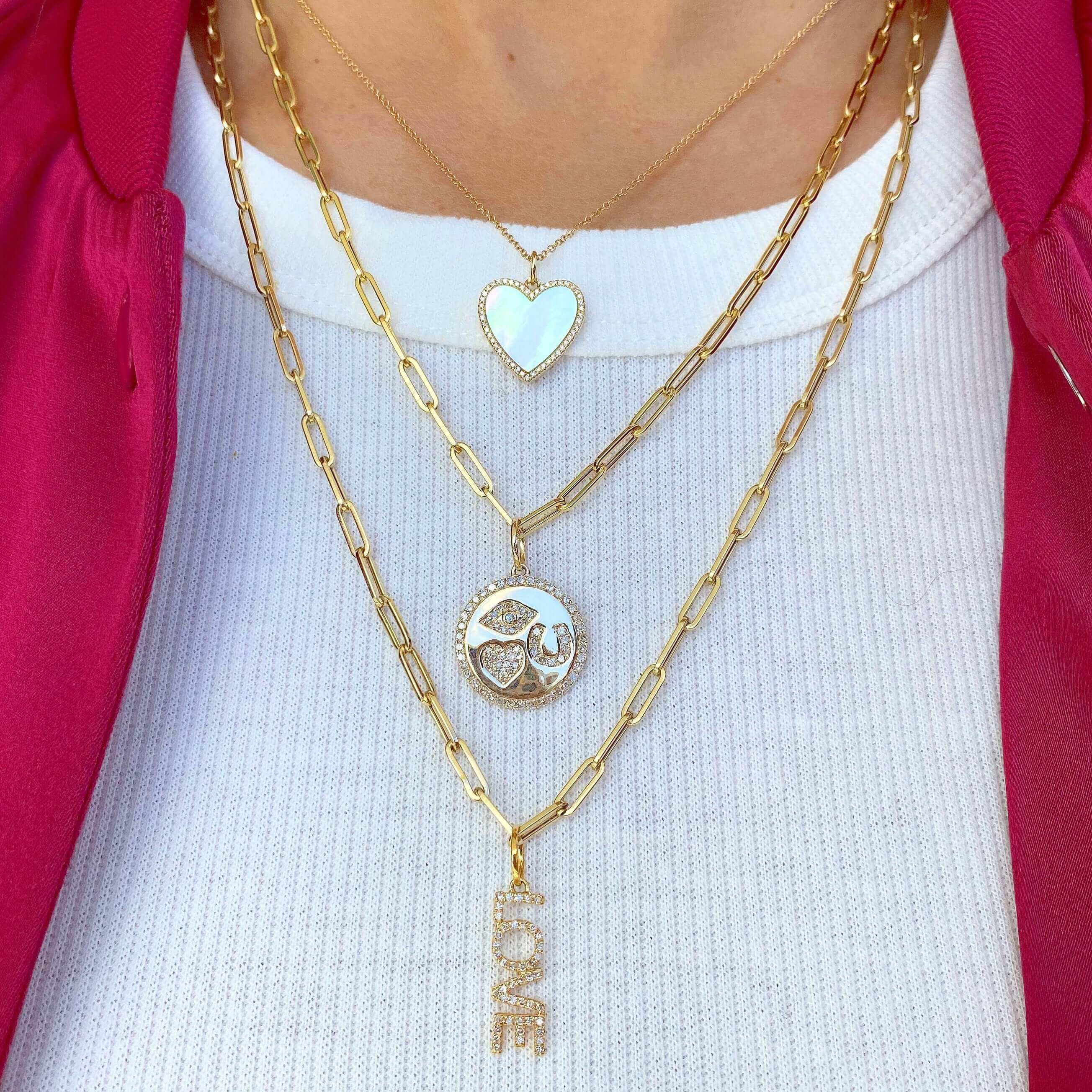 Layered necklaces with 14K Gold Diamond Pave Eye Heart U Coin Charm, heart pendant, and “LOVE” charm on gold chains.