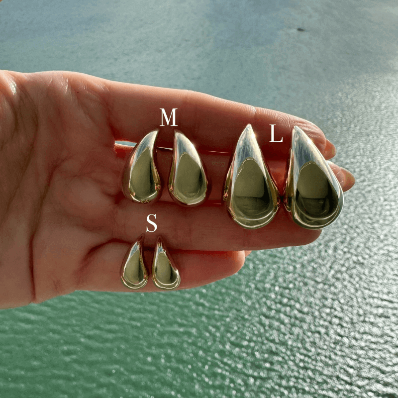 Hand holding three sizes of teardrop-shaped silver earrings against a serene waterfront backdrop