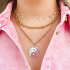 Layered gold necklaces featuring yin-yang pendant and heart charm, worn on pink shirt